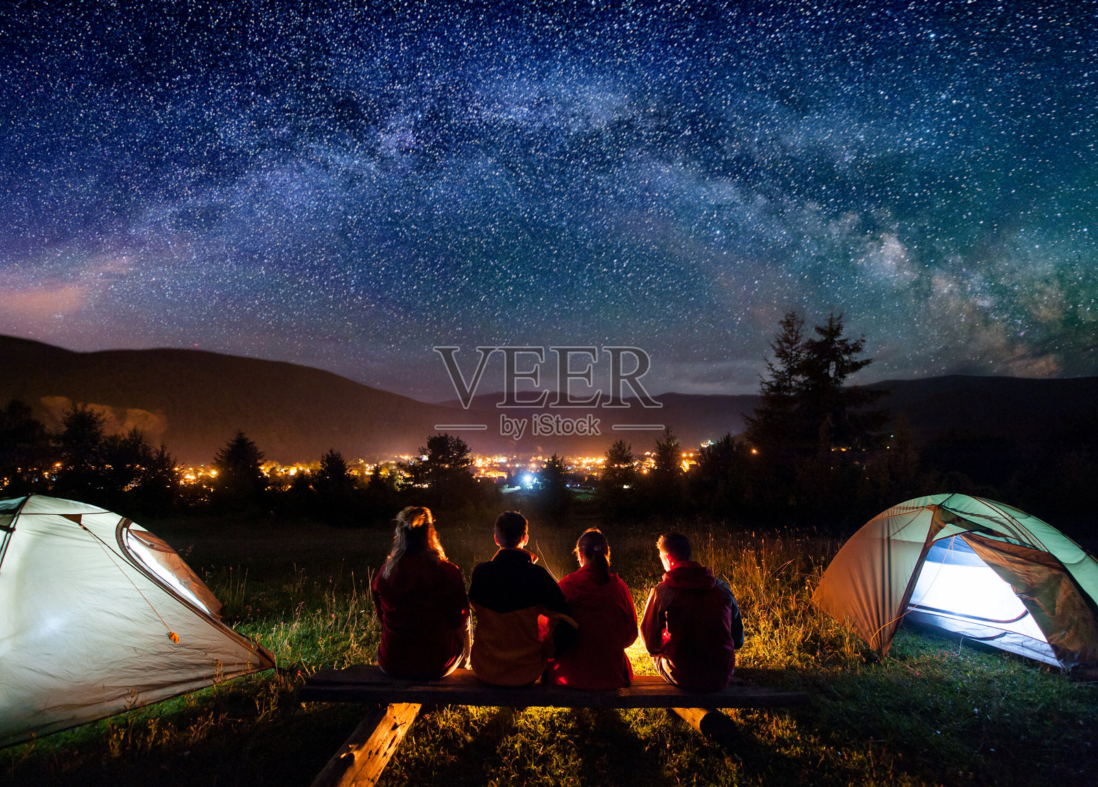4个人的剪影坐在一个由原木制成的长凳上，一起看火在营地和帐篷旁边的夜晚。背景是星空、银河、群山和发光的小镇。后视照片摄影图片