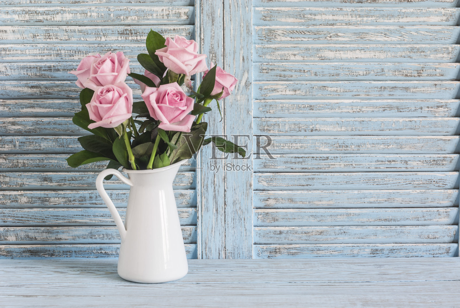 粉红色的玫瑰在白色搪瓷壶在蓝色的乡村背景。文本的空闲空间。复古风格照片摄影图片