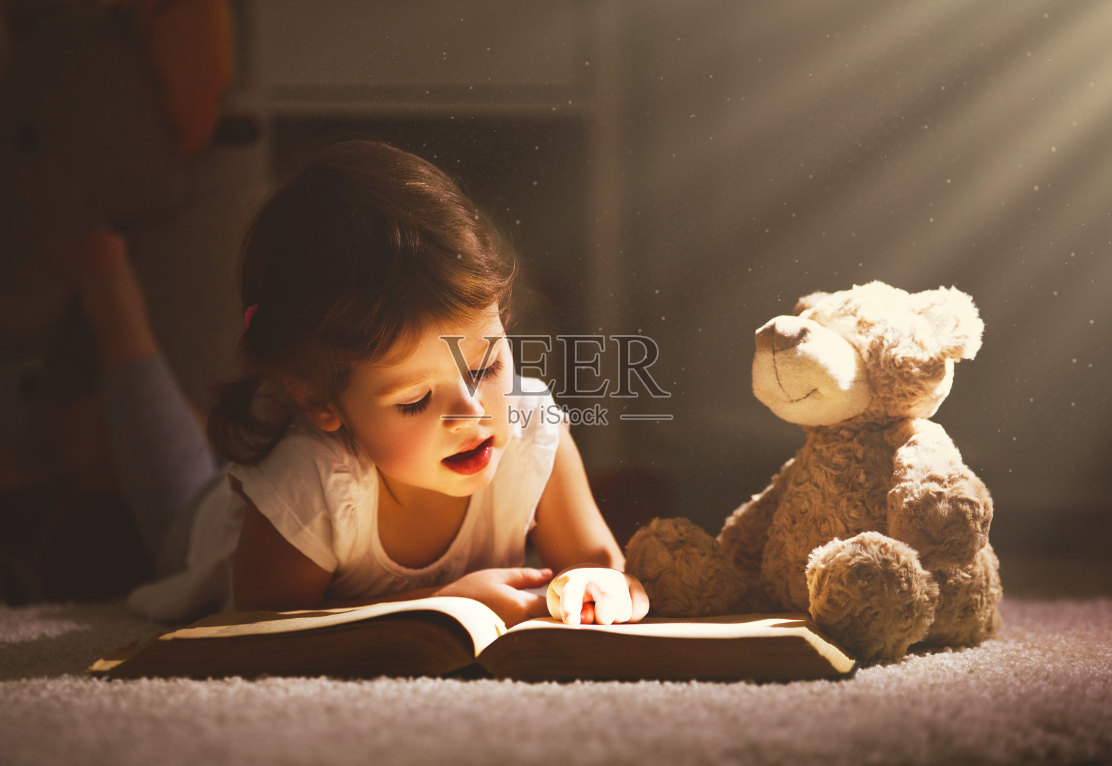 一个小女孩晚上在黑暗中和一只玩具熊一起看书照片摄影图片