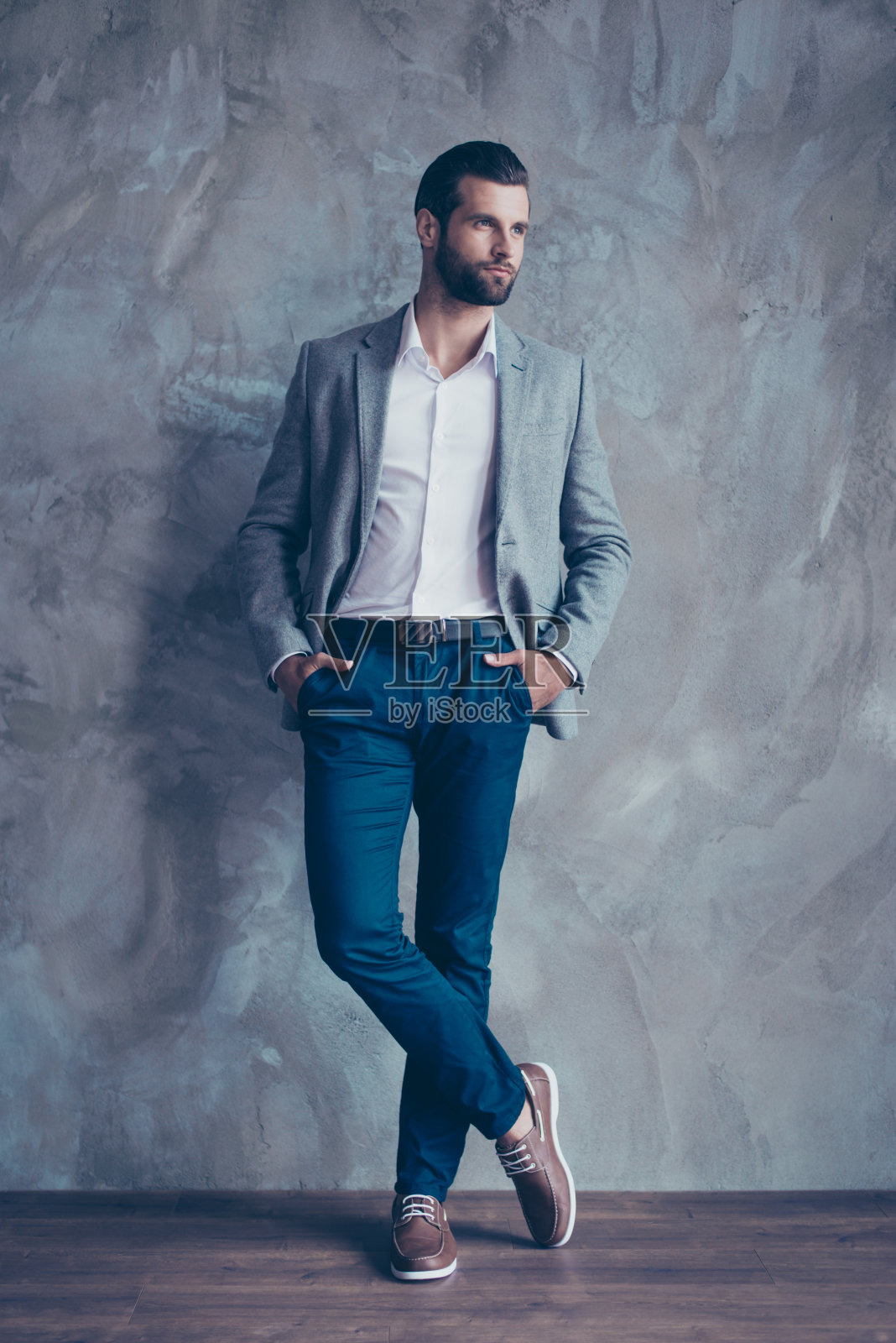 全尺寸肖像时尚的年轻胡子男子站在灰色的混凝土背景。他穿着西装，双腿交叉站着照片摄影图片