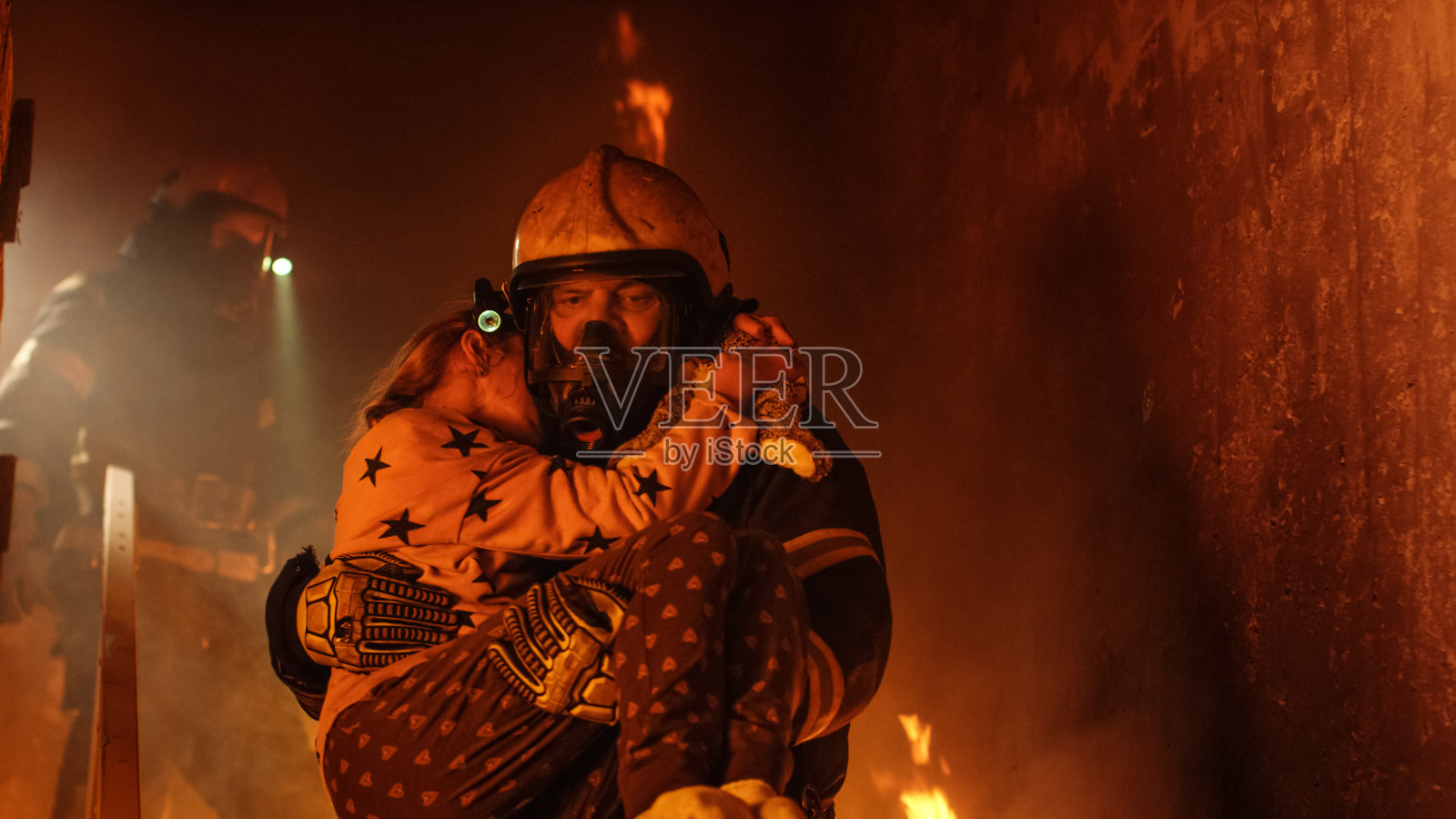勇敢的消防队员走下燃烧的建筑物的楼梯，将获救的女孩抱在怀里。开火，背景中有一名消防员。照片摄影图片