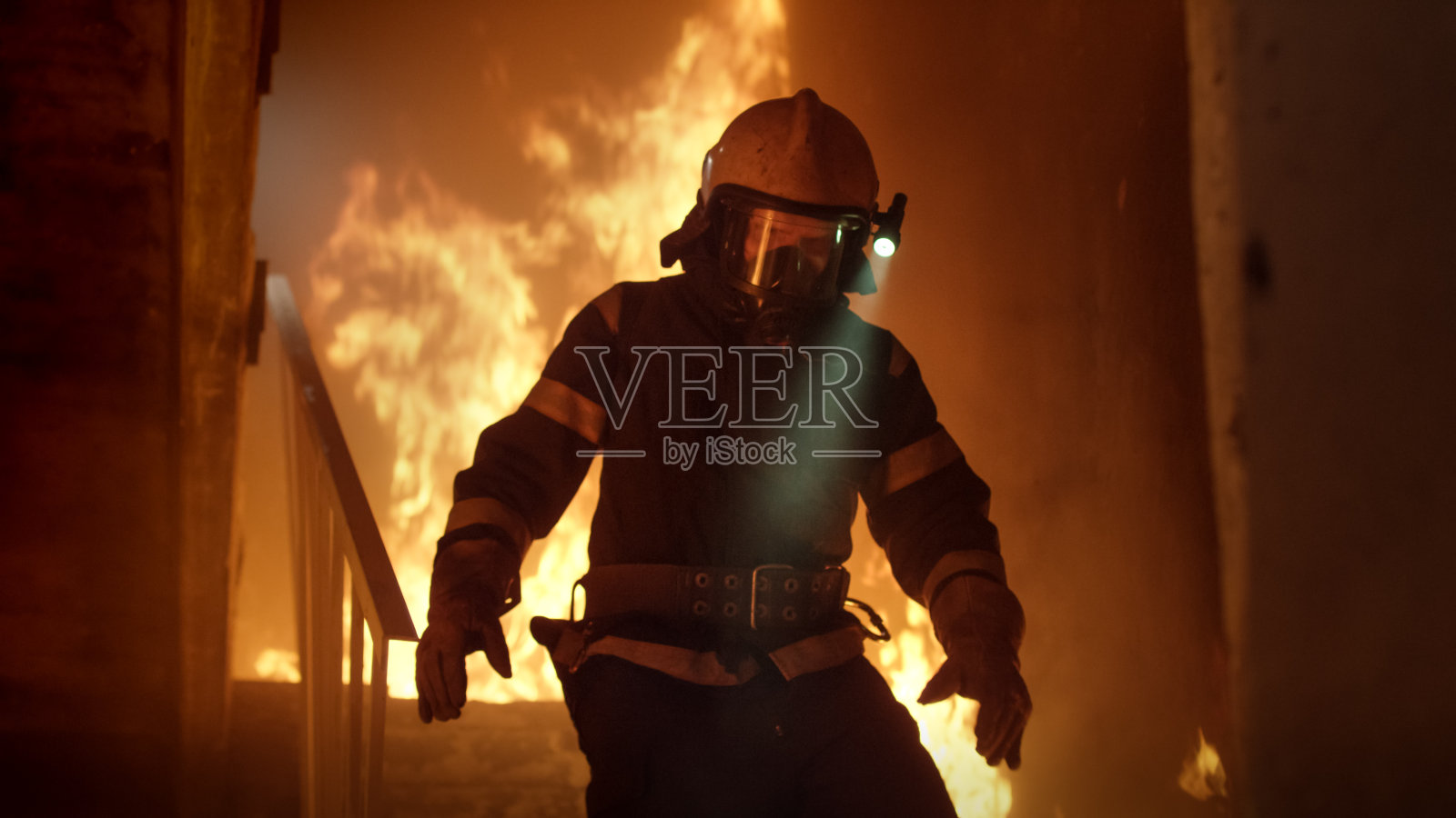 勇敢的消防员在头盔上打开手电筒跑下燃烧的楼梯。火是激烈的。照片摄影图片
