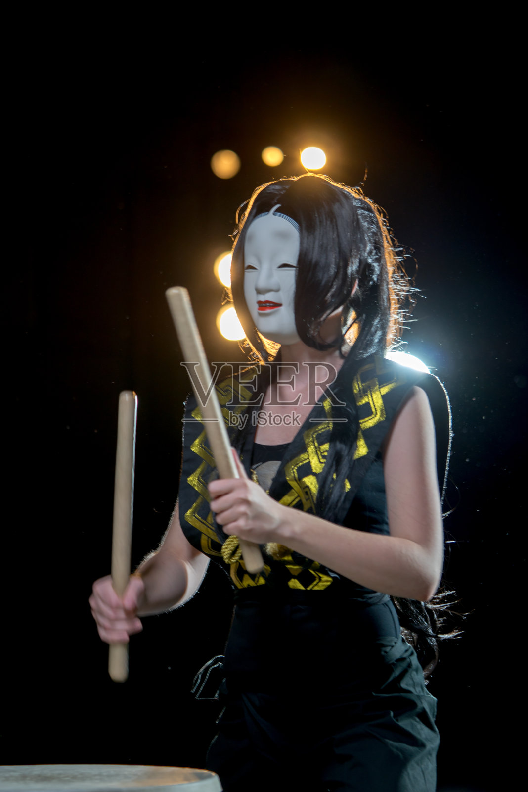 鼓手女孩在白色的恶魔面具与鼓槌，工作室音乐会拍摄在一个黑暗的背景和天窗灯。照片摄影图片
