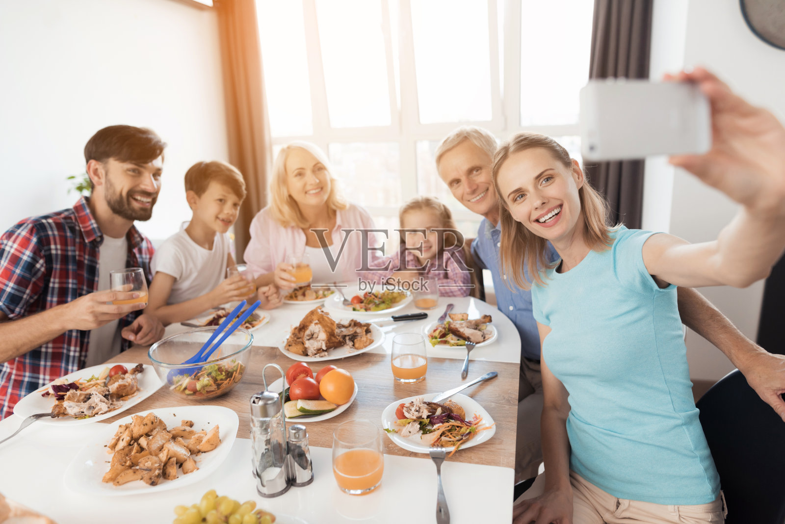 一名身穿蓝色t恤的女子在白色智能手机上自拍。她的家人坐在她身后的节日餐桌旁照片摄影图片
