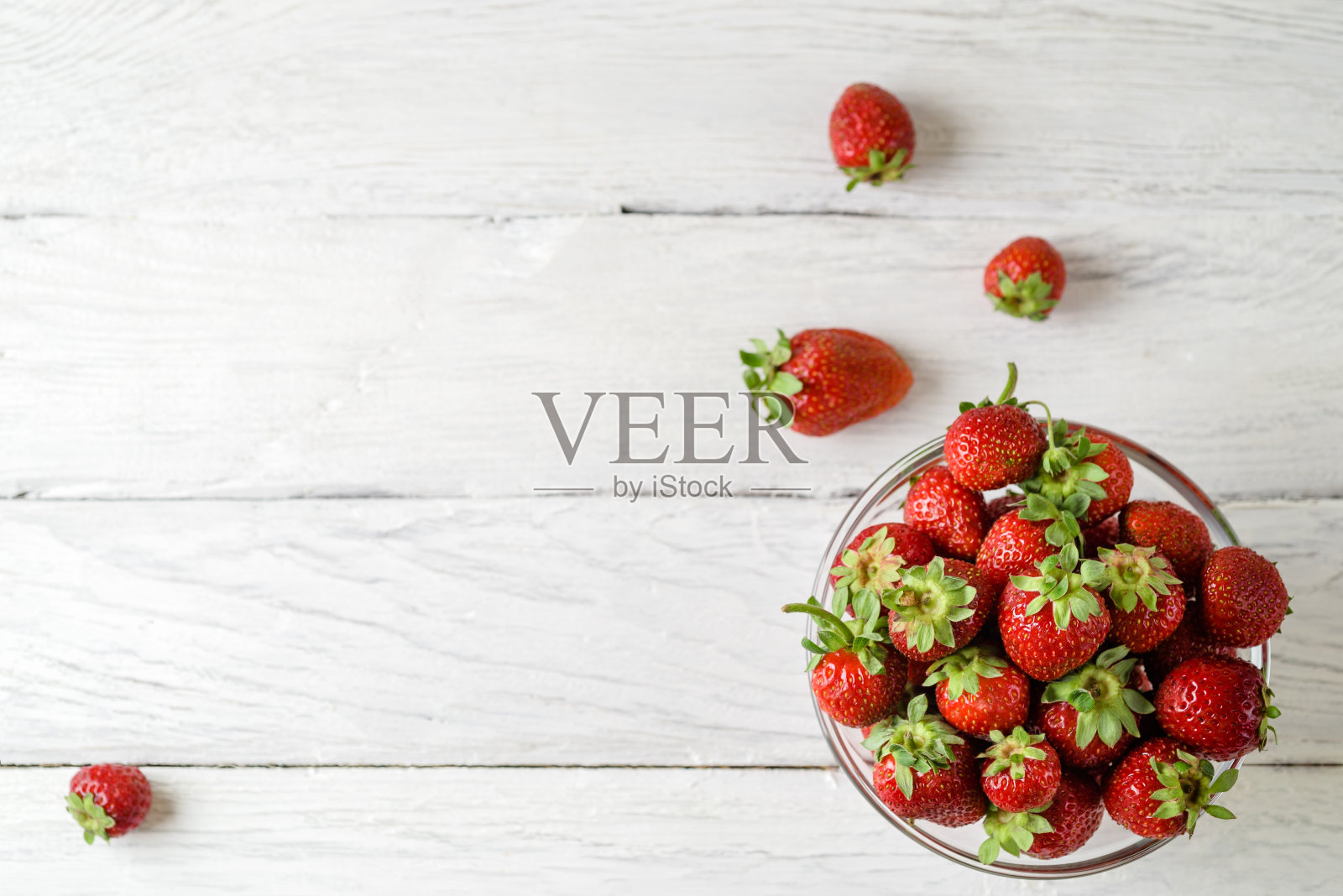 俯视图的照片，一个玻璃盘子与成熟的红色草莓在白色木制桌子上。桌上散落着四颗草莓。照片摄影图片
