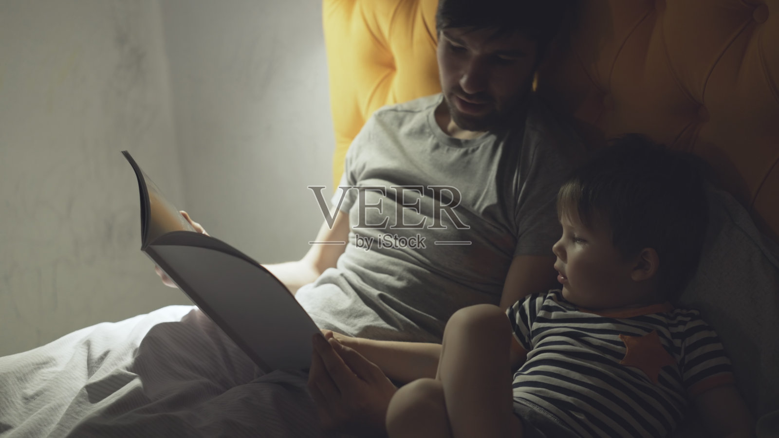 晚上睡觉前，年轻的父亲和小儿子躺在家里的床上看童话书照片摄影图片