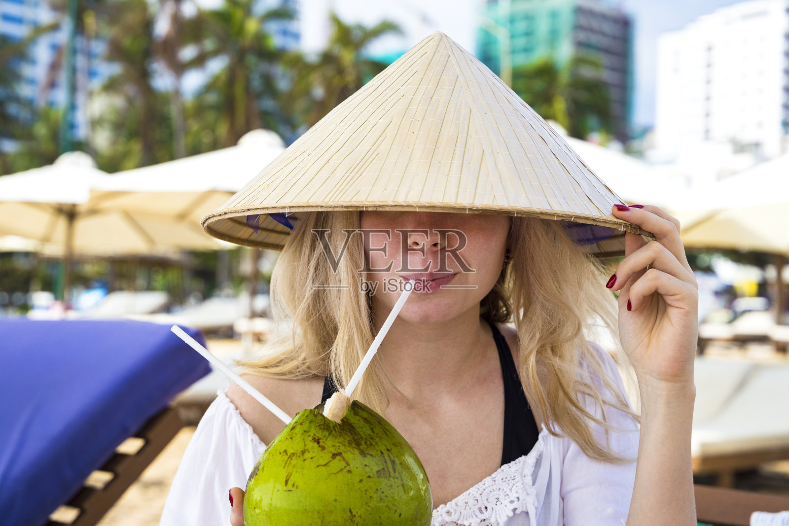 年轻美丽的女孩坐在海边的沙滩上，头戴亚洲传统的锥形帽子，喝着椰子汁照片摄影图片