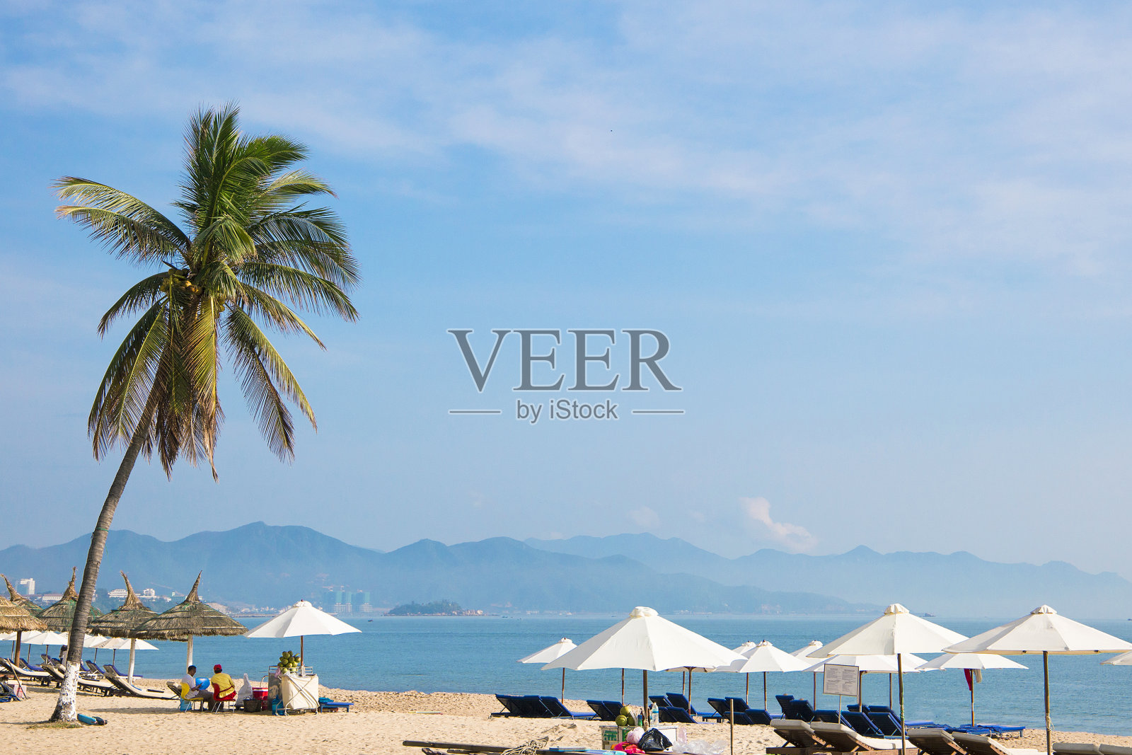 沙滩上有棕榈树，白色的伞，沙滩椅在蓝色的海边，以山和城市为背景照片摄影图片
