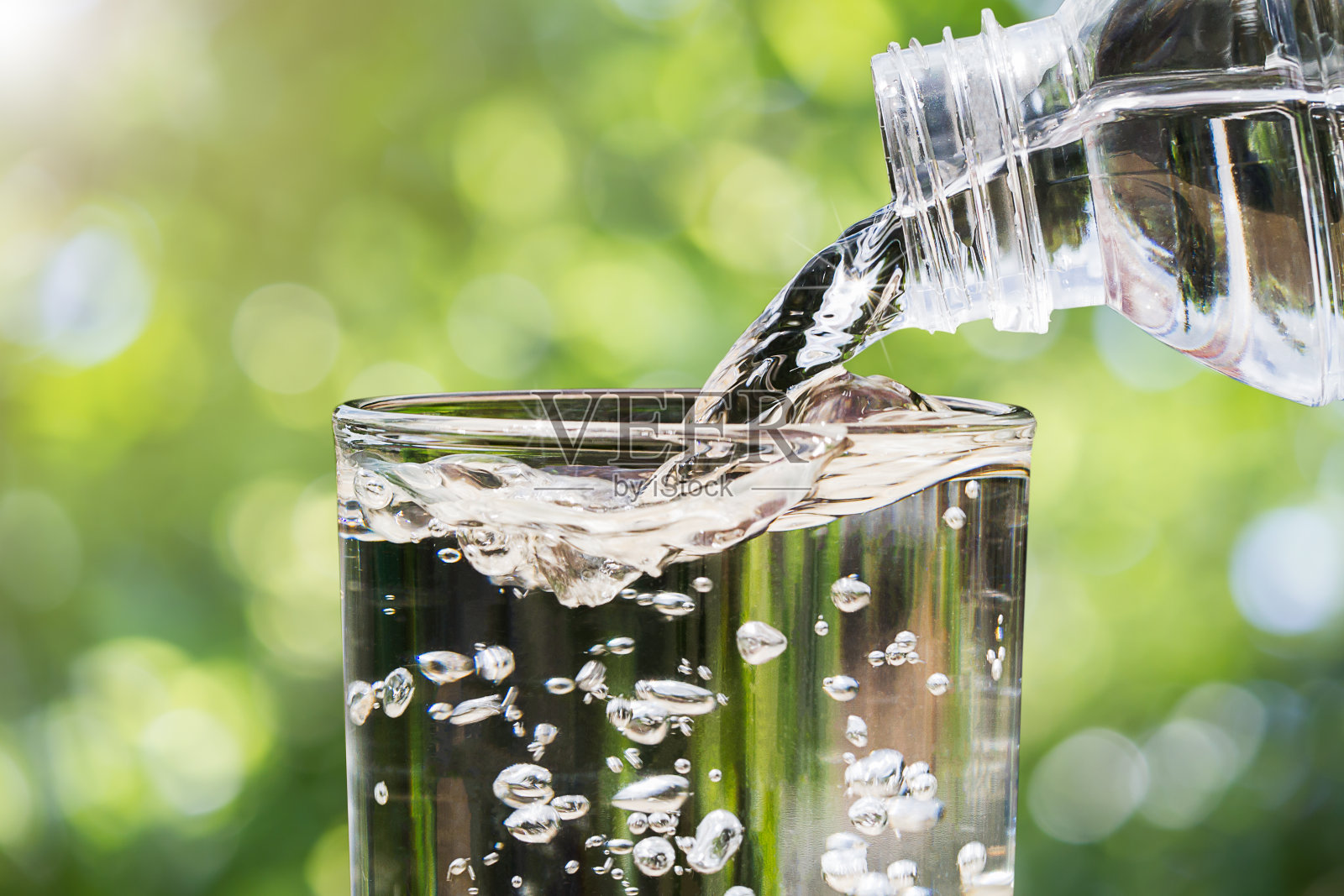 特写水从饮用水瓶流入玻璃上模糊的绿色自然散景背景，健康饮用干净的水的概念照片摄影图片