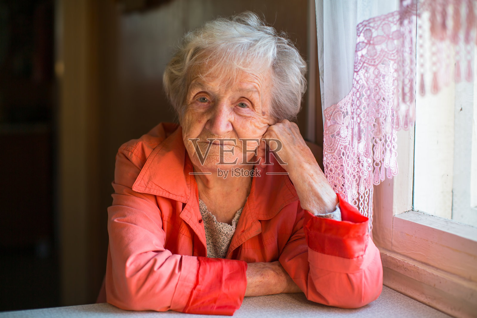 穿着红色夹克的老妇人坐在家里的桌子旁。照片摄影图片