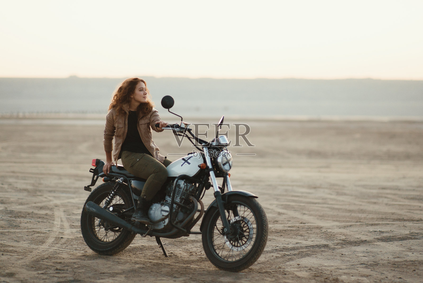 年轻美丽的女人坐在她的老咖啡赛车摩托车在沙漠日落或日出照片摄影图片