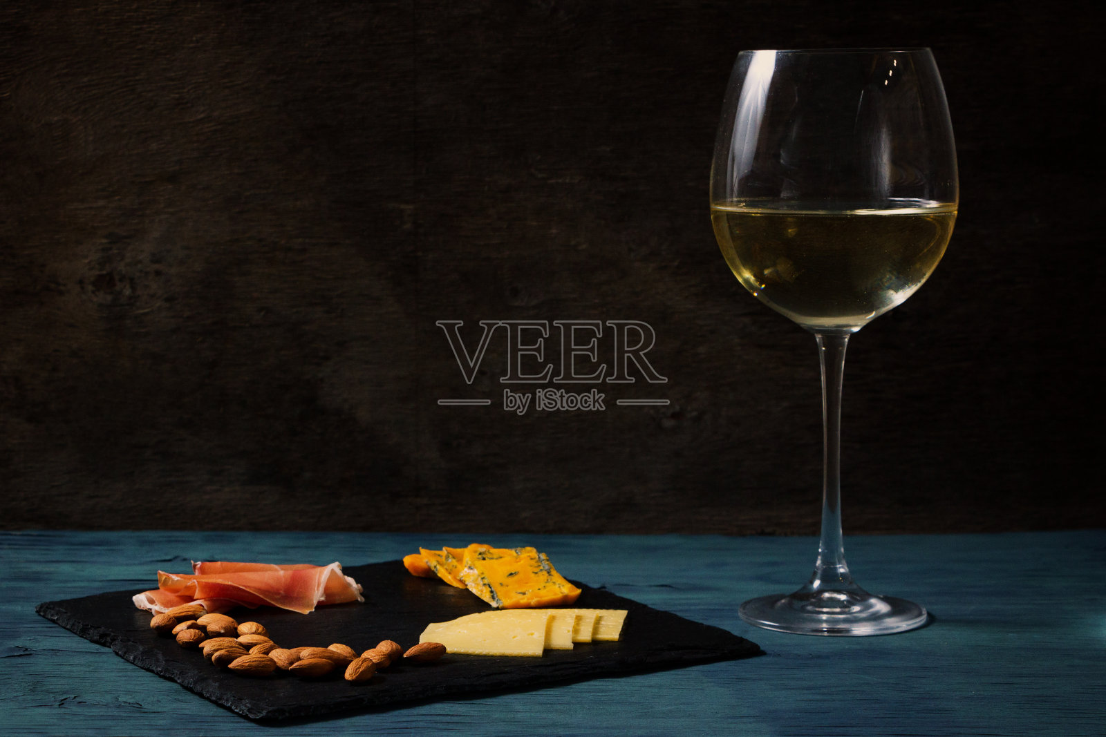 石板托盘上放着零食，蓝色和黑色背景上放着一杯白葡萄酒。奶酪、坚果、火腿。作者处理,copy-space照片摄影图片