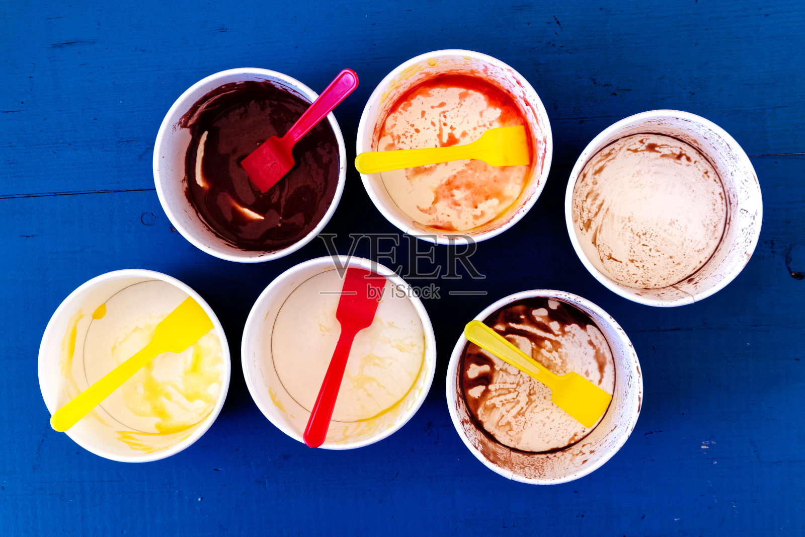 6个凌乱的空冰淇淋纸杯，黄色和红色的塑料勺子从上面被隔离在蓝色油漆的木头上。照片摄影图片