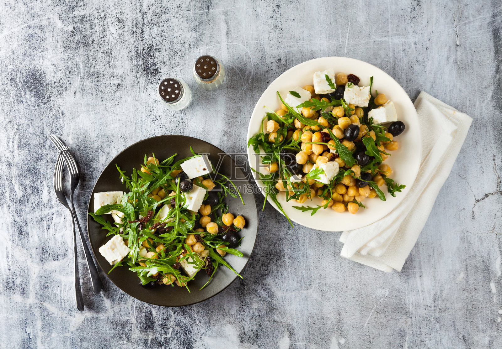 两盘蔬菜沙拉，配鹰嘴豆、芝麻菜、羊乳酪或豆腐和黑橄榄，背景是天然的石头。健康饮食照片摄影图片