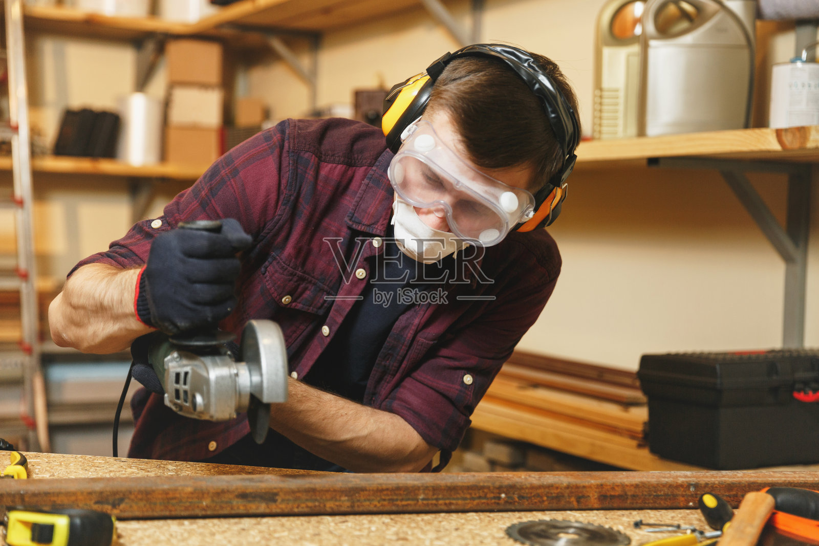 白人青年男子在格子衬衫，黑色t恤，隔音耳机，防护面具工作在木工车间的木桌地方与不同的工具，锯铁与电锯。照片摄影图片