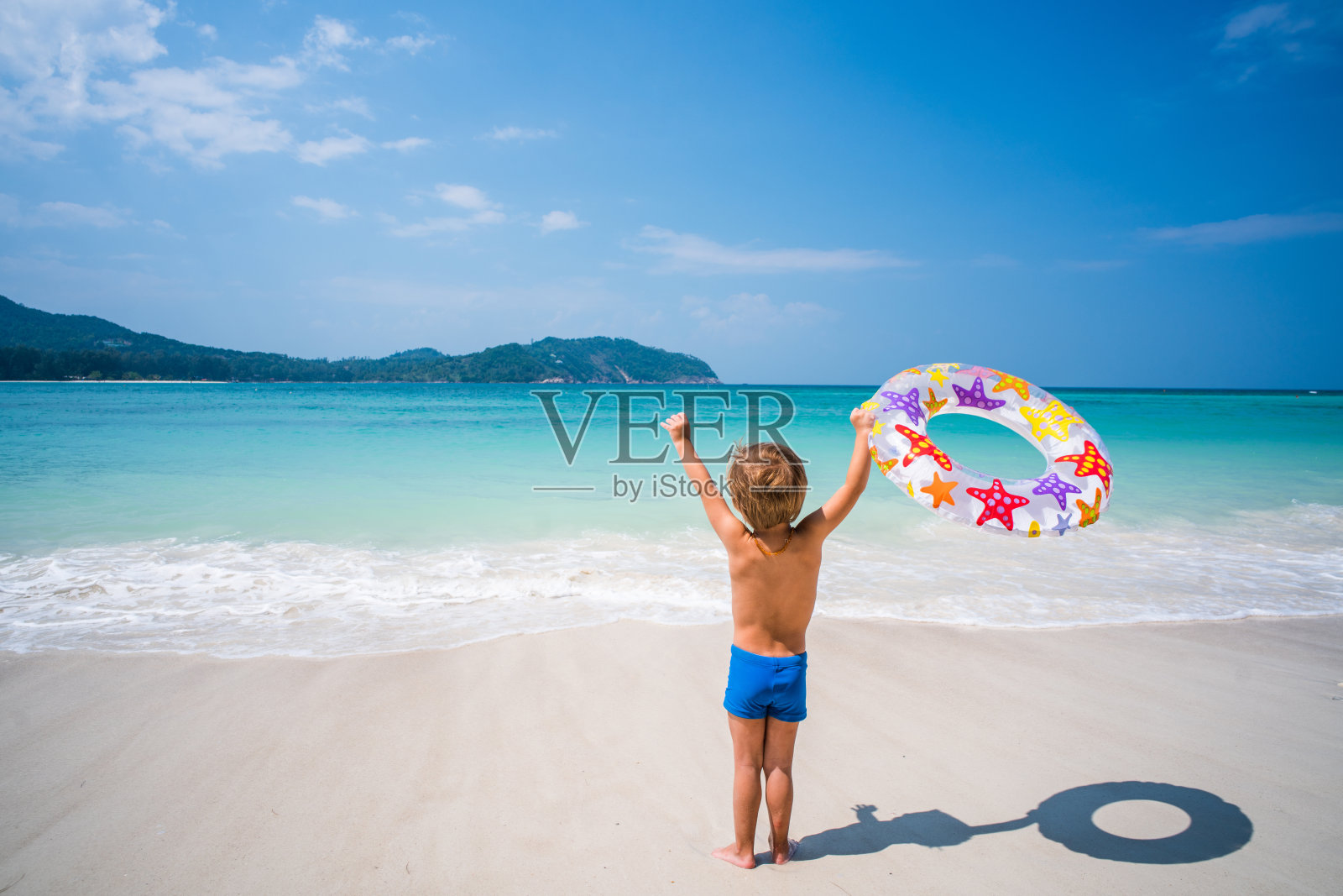 无忧无虑的男孩的后视图举起手臂在夏天的一天在海滩上。照片摄影图片