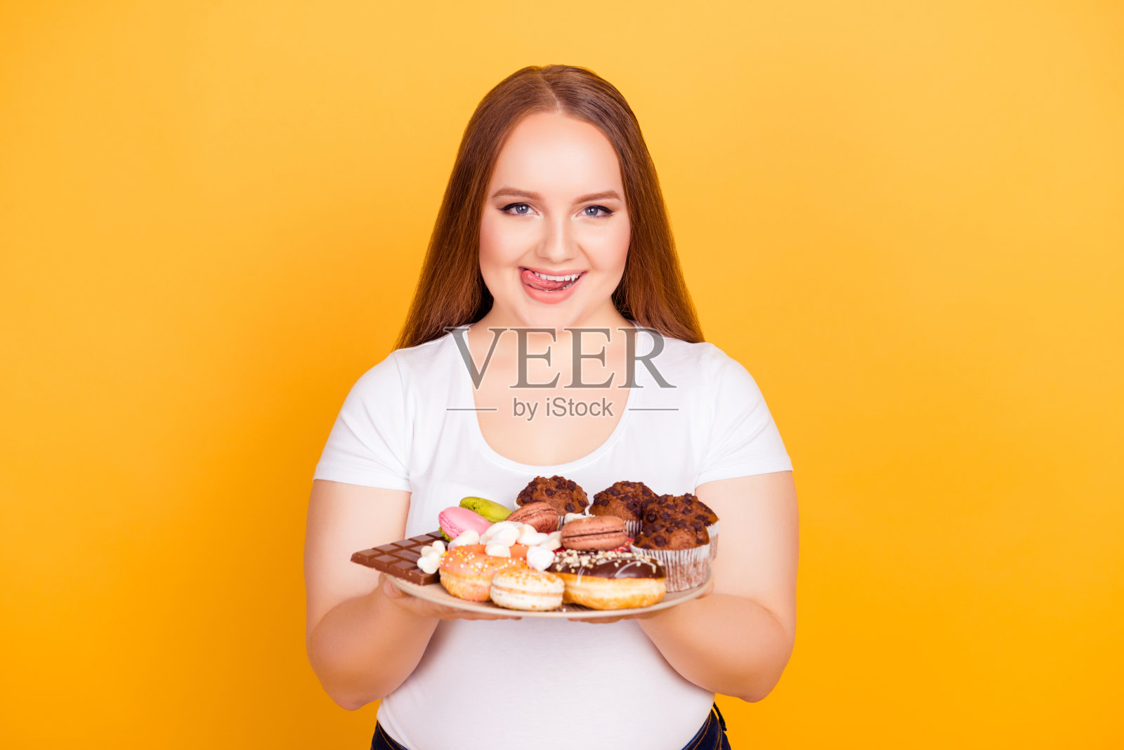 肖像快乐快乐饥饿的年轻女子穿着白色t恤，她是拿着一个盘子与高热量的产品和舔她的嘴唇，在明亮的黄色背景，照顾，身体照片摄影图片