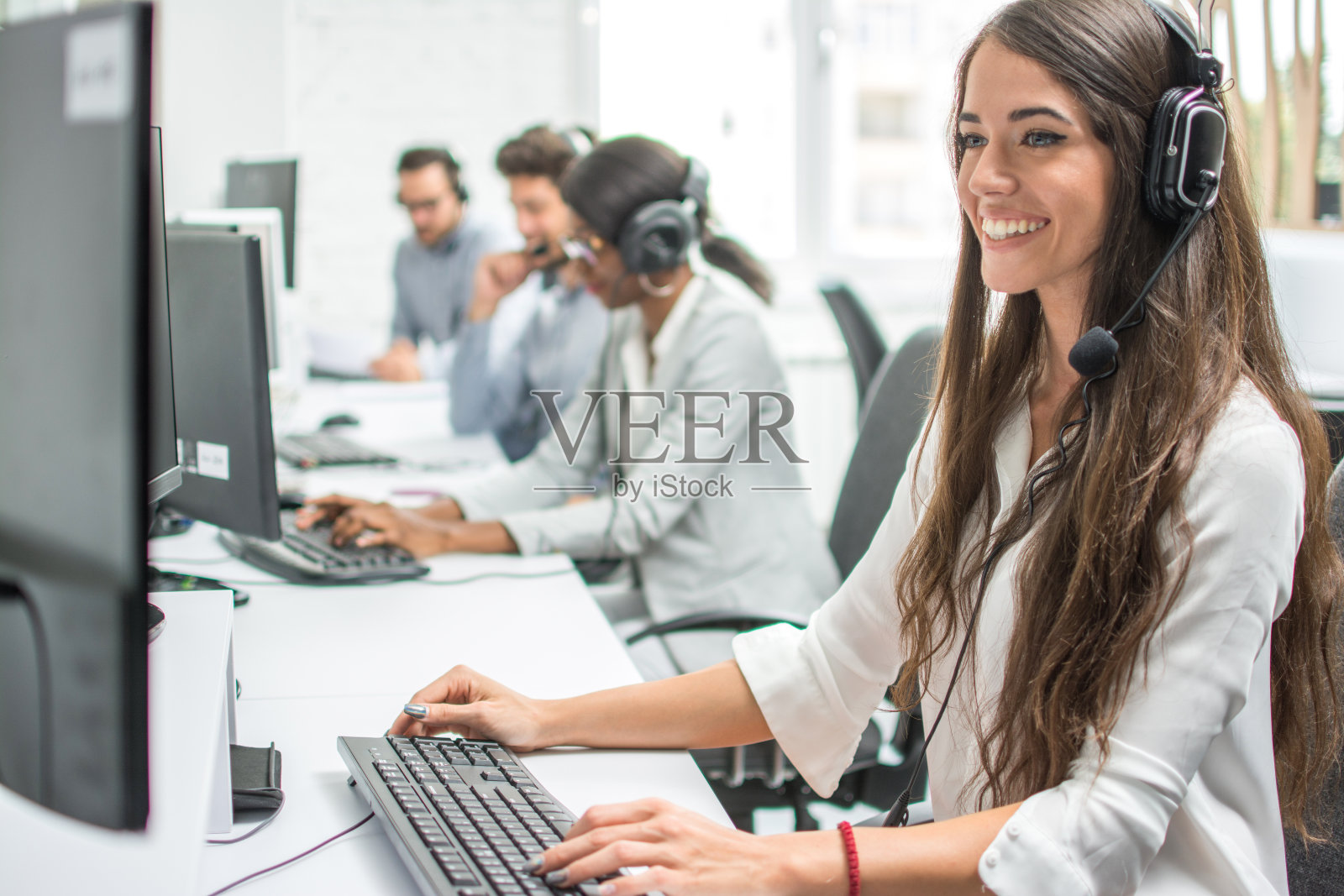 年轻的微笑接线员女性代理人与耳机工作在一个呼叫中心。照片摄影图片