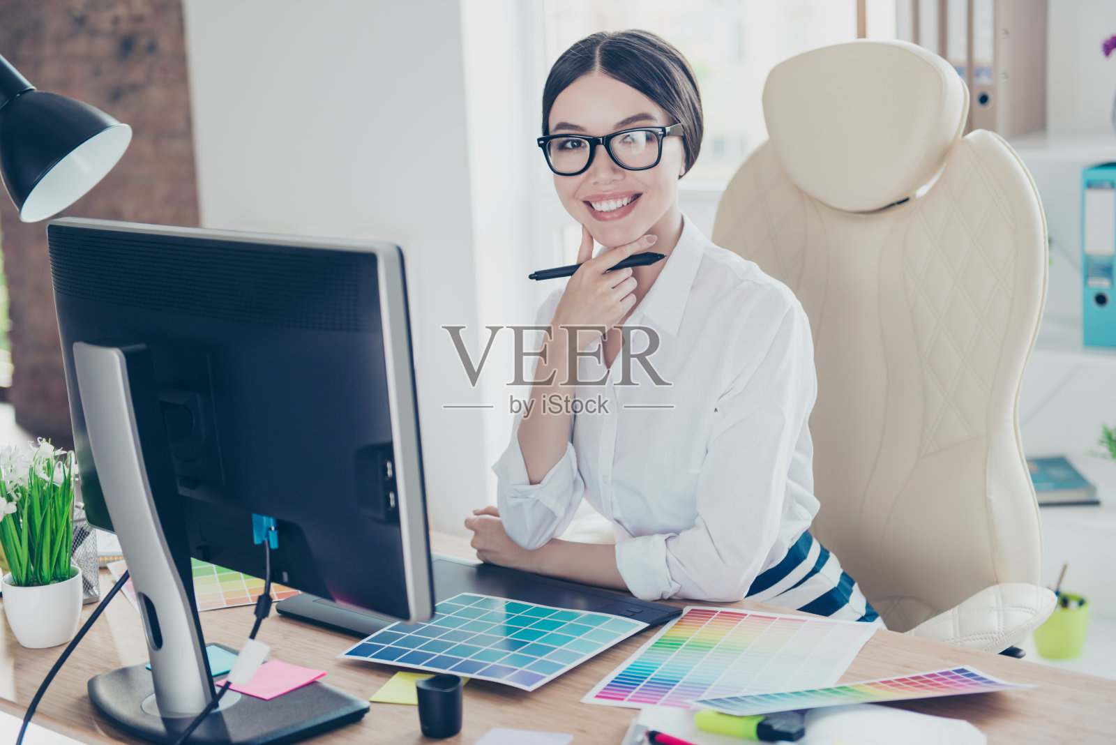 成功的有吸引力的商务女性设计师坐在她的工作岗位上手写笔，穿着正式，戴着眼镜，这样一个可爱的聪明的专业人士照片摄影图片