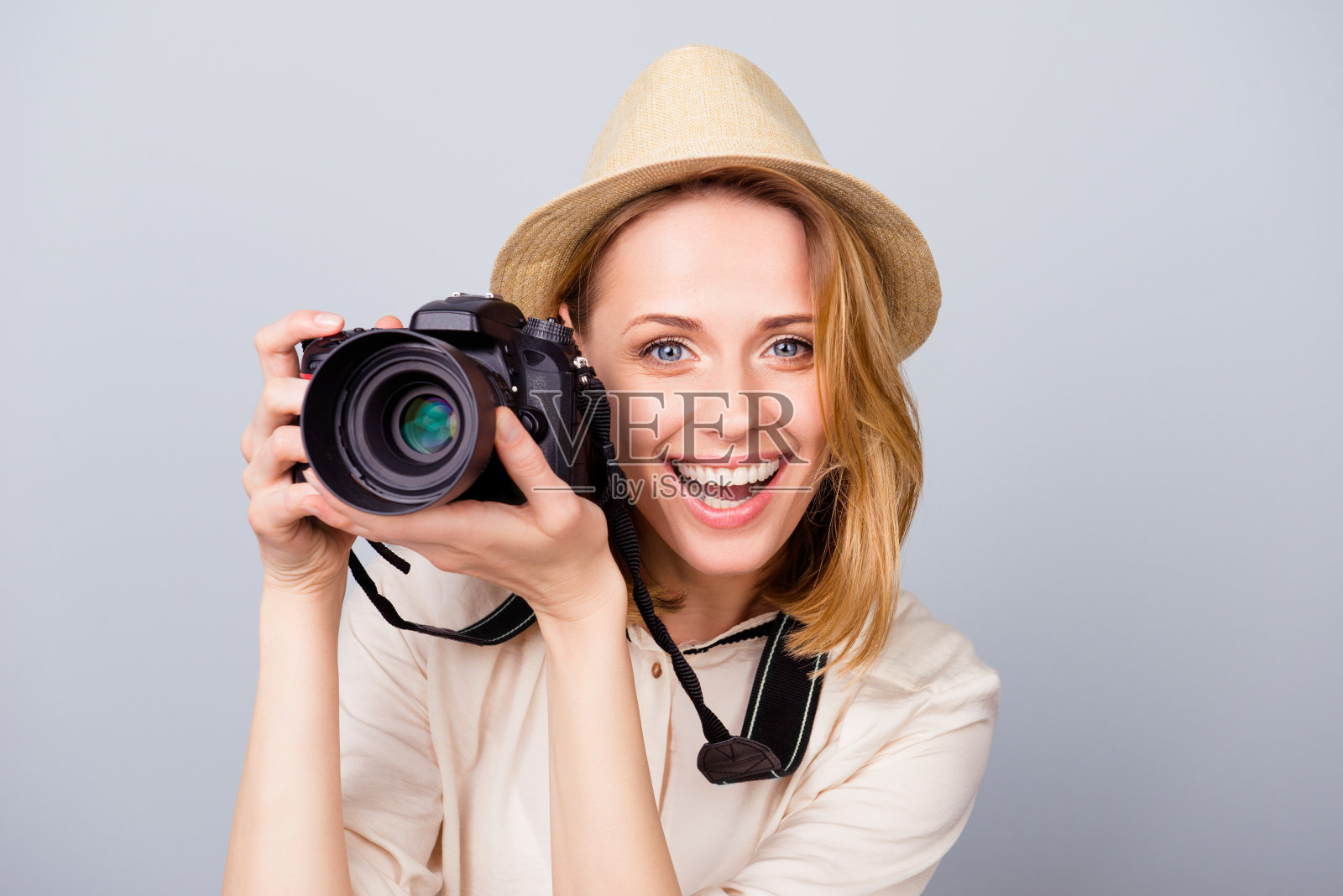 年轻开朗的金发女摄影师在淡蓝色的背景上微笑着。她很兴奋，拿着相机，戴着帽子照片摄影图片