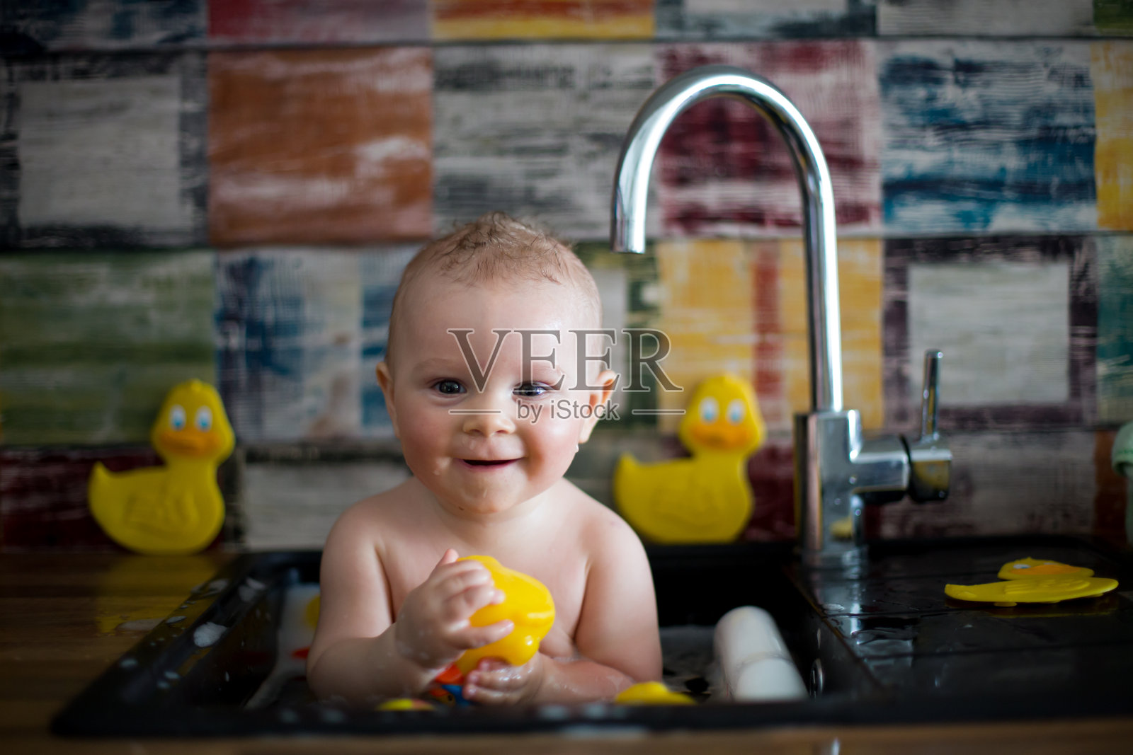 可爱的婴儿在厨房的水槽洗澡。孩子在阳光明媚的厨房里和橡皮鸭一起玩泡沫和肥皂泡，小男孩在洗澡照片摄影图片