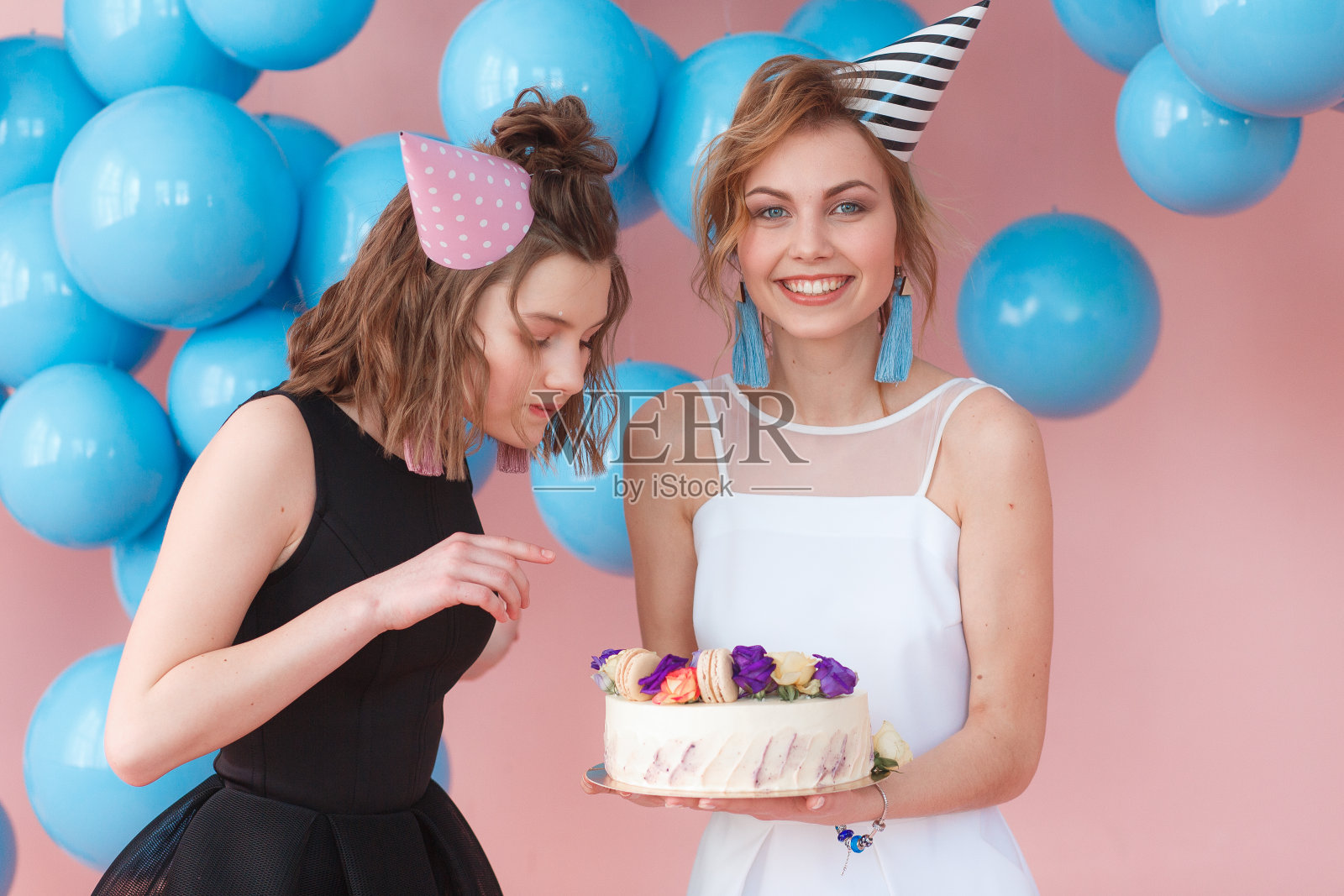 两个戴派对帽的少女拿着蛋糕。孤立在粉红色背景和蓝色气球照片摄影图片