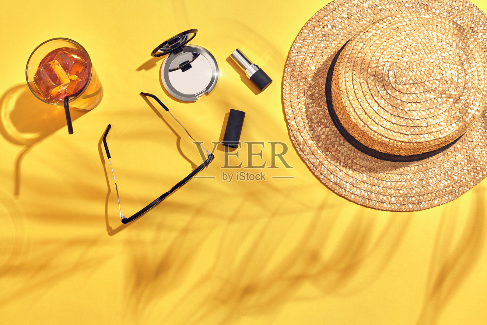 草海滩妇女的帽子，饮料与冰在一个玻璃杯和太阳眼镜在黄色的背景与棕榈叶的阴影照片摄影图片