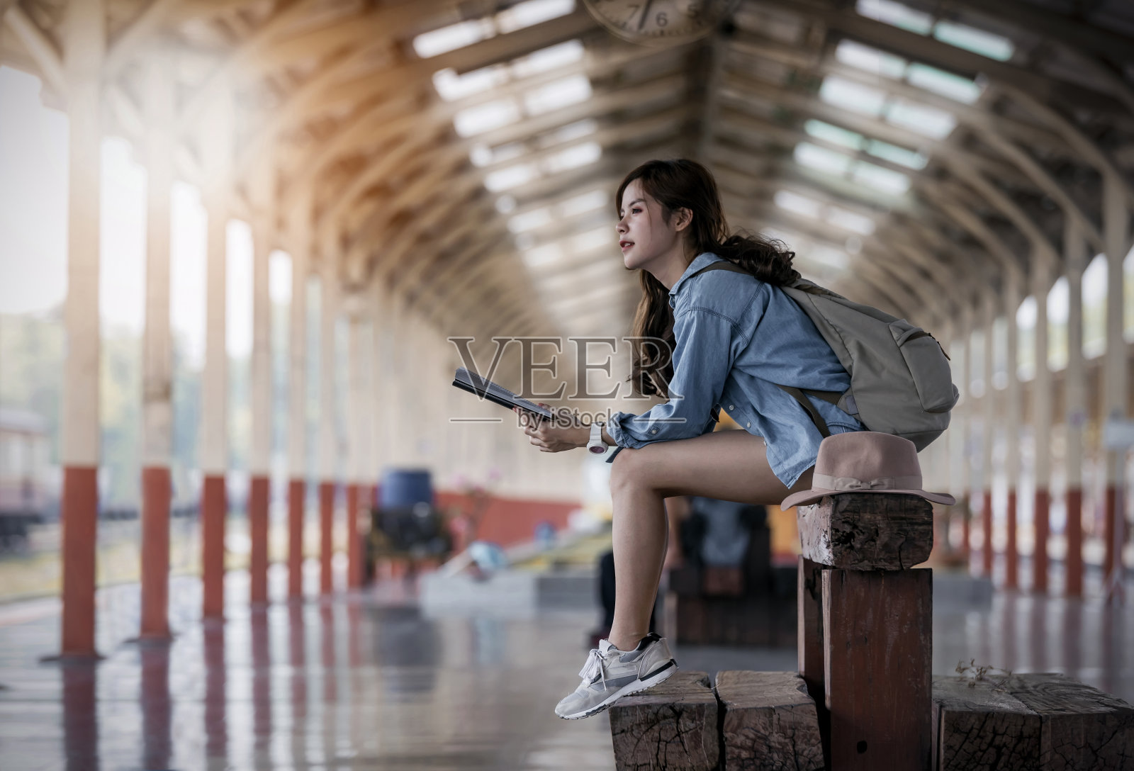 穿着蓝色牛仔裤，拿着书的女旅客坐在火车站月台的木凳上照片摄影图片