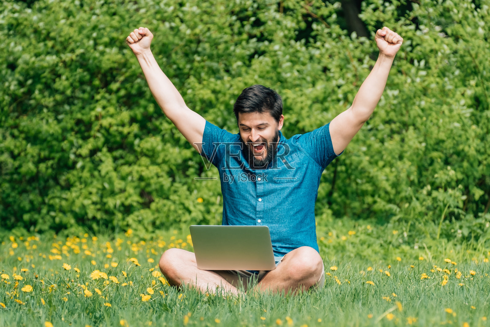 微笑的年轻人坐在绿色的草地上，举起他的双手和笔记本电脑在他的交叉腿上照片摄影图片