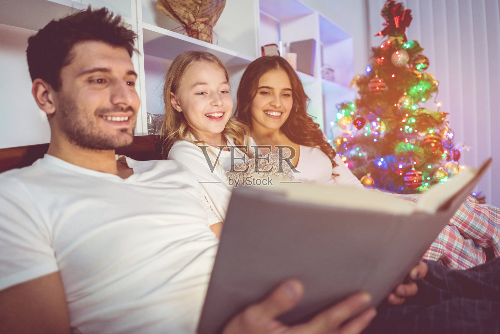 幸福的一家人在圣诞树旁的床上看这本书。晚上的时间照片摄影图片