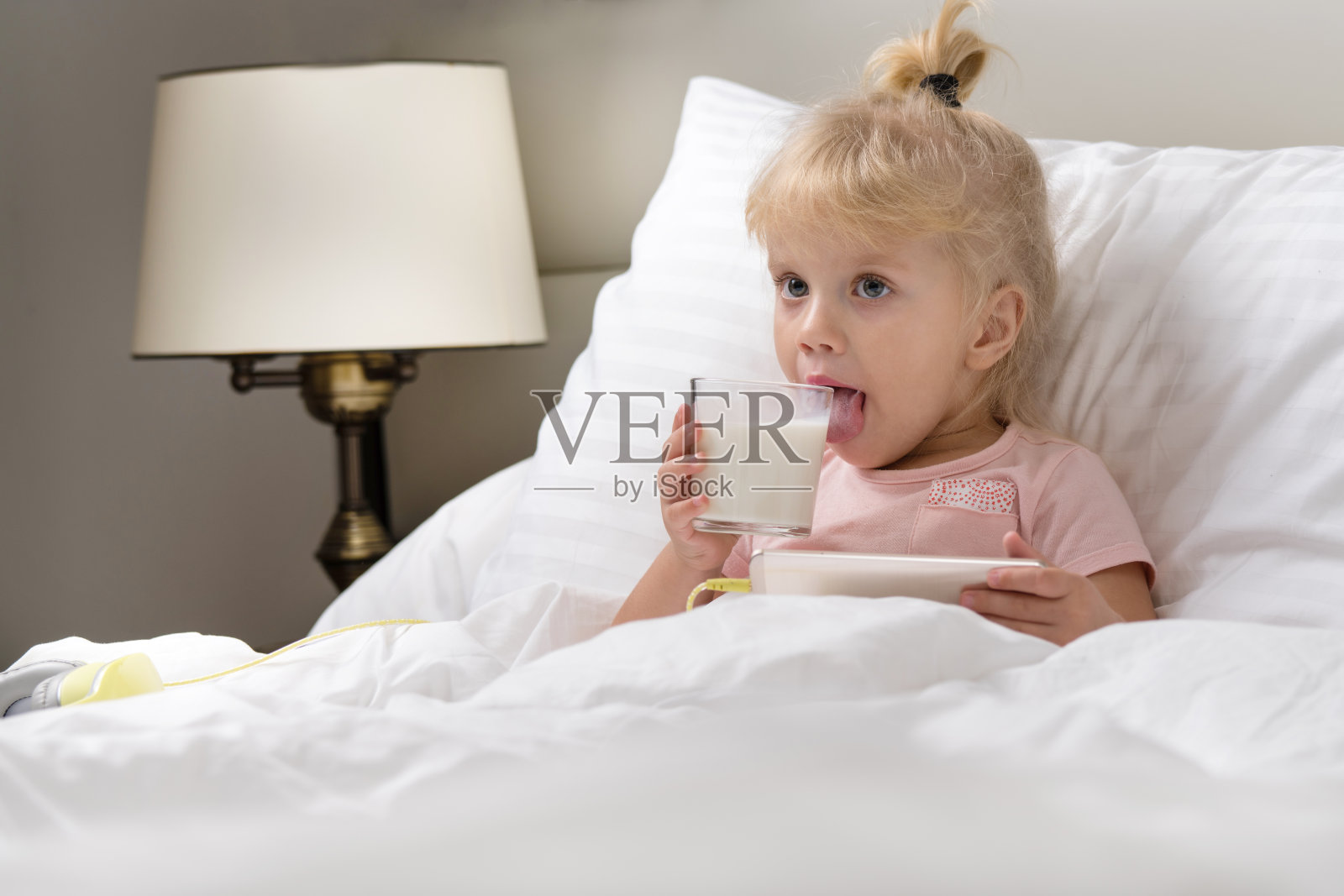 小女婴躺在白色的床上，用智能手机玩着，早上喝着牛奶照片摄影图片