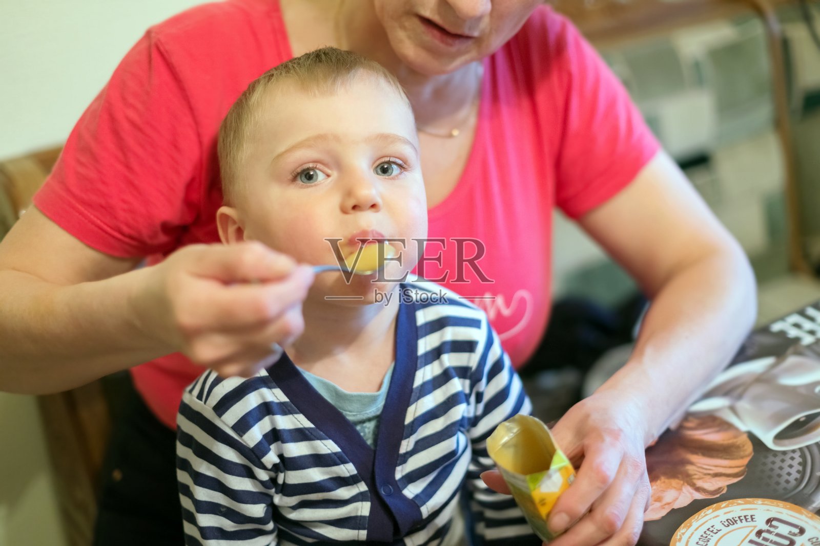 一个笑着的小男孩，1.5岁，坐在女人的膝盖上，用勺子吃水果泥。照片摄影图片