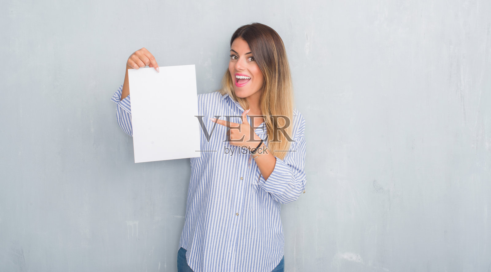 年轻的成年妇女在灰色的垃圾墙拿着空白的纸非常高兴的指着手和手指照片摄影图片