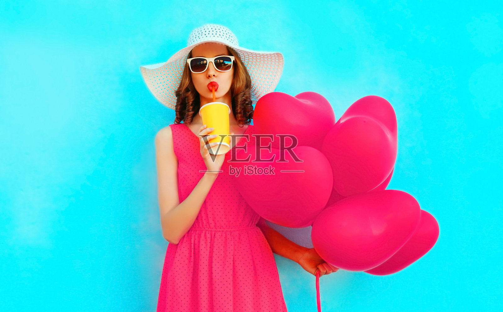 肖像美丽的女人喝果汁与粉红色的心形气球在彩色的蓝色背景，戴着夏季草帽照片摄影图片