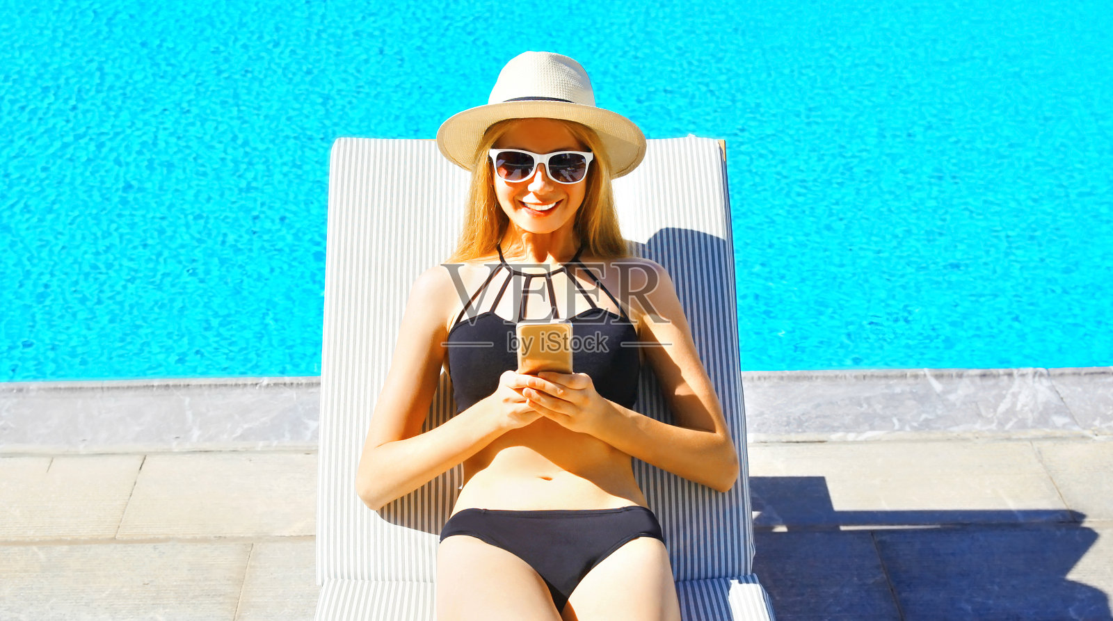 暑假概念——快乐的年轻女子躺在帆布躺椅上用智能手机看蓝色的游泳池照片摄影图片