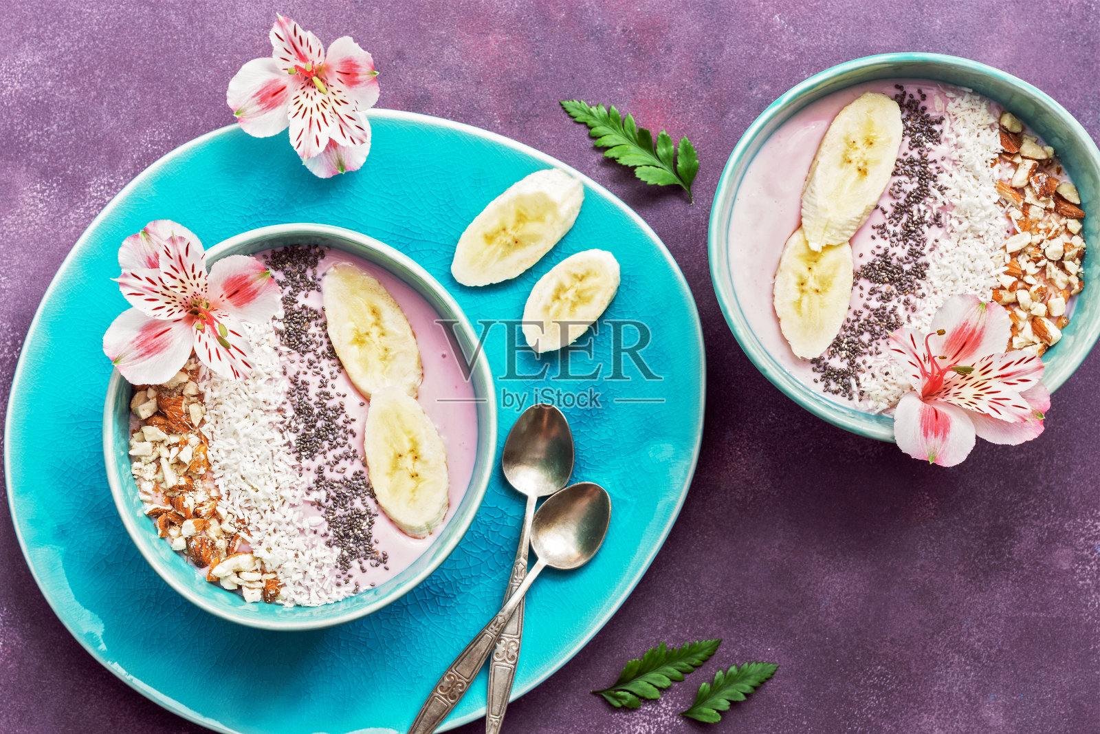 新鲜健康的早餐-酸奶香蕉，杏仁，椰子片，奇异子在一个蓝色的碗装饰花在紫色的背景。俯视图照片摄影图片