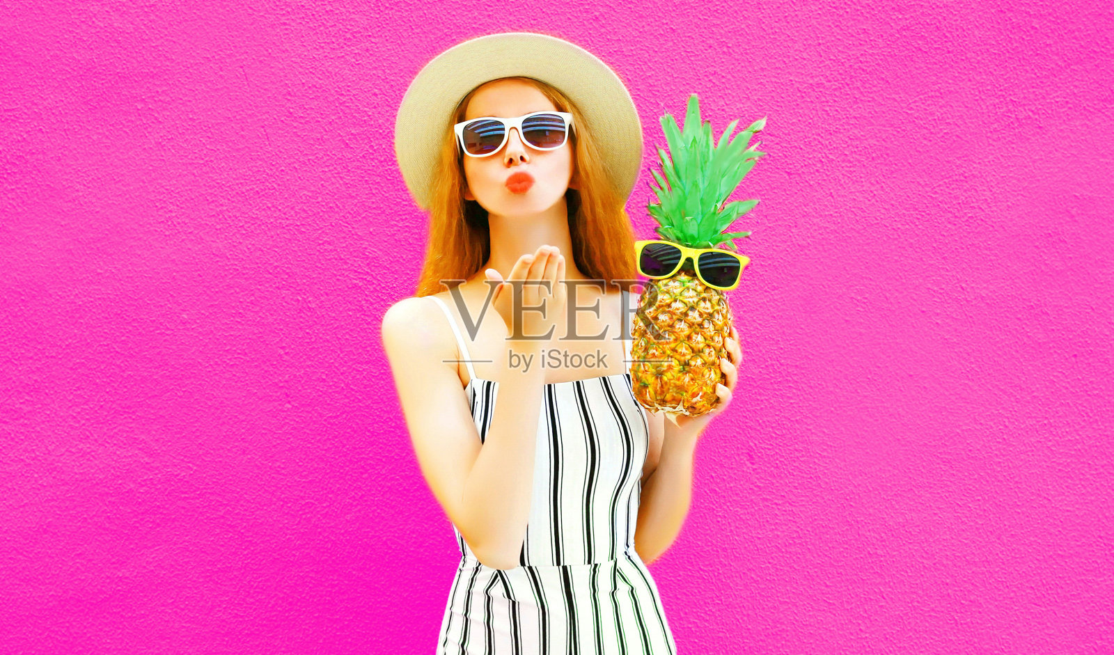 肖像时尚的女人发送甜蜜的飞吻与菠萝在夏季圆形草帽，白色条纹连衣裤在彩色的粉红色墙壁背景照片摄影图片
