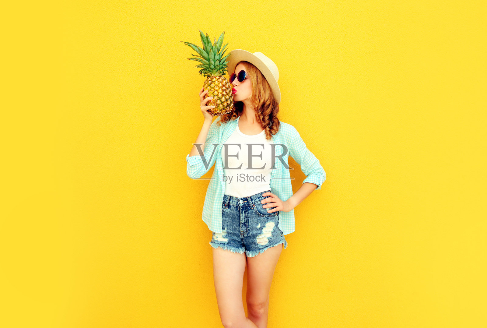 美丽的年轻女子亲吻菠萝在夏天草帽，短裤上彩色的黄色背景照片摄影图片