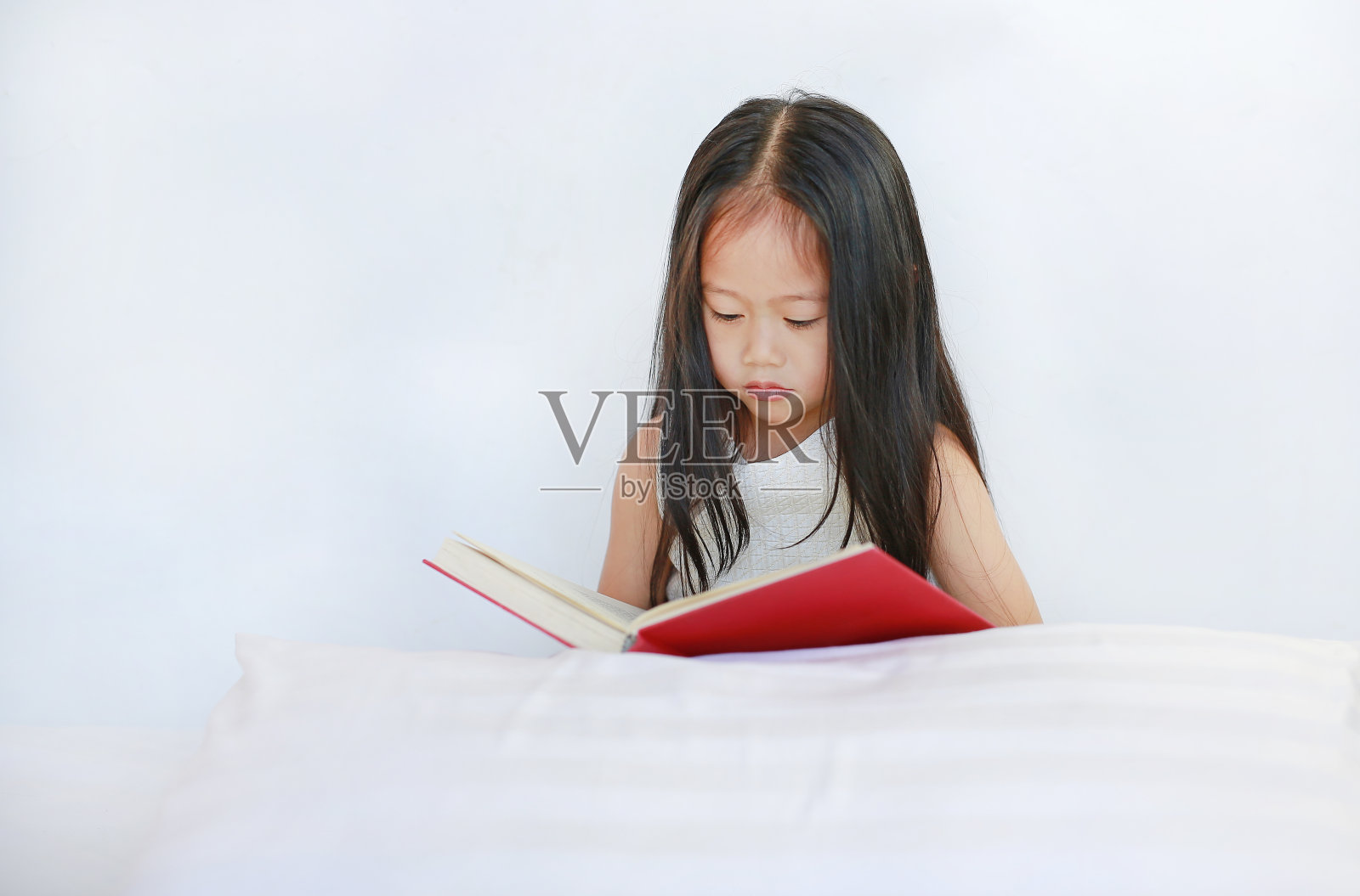 美丽的亚洲小女孩读精装书与枕头躺在白色的背景。照片摄影图片