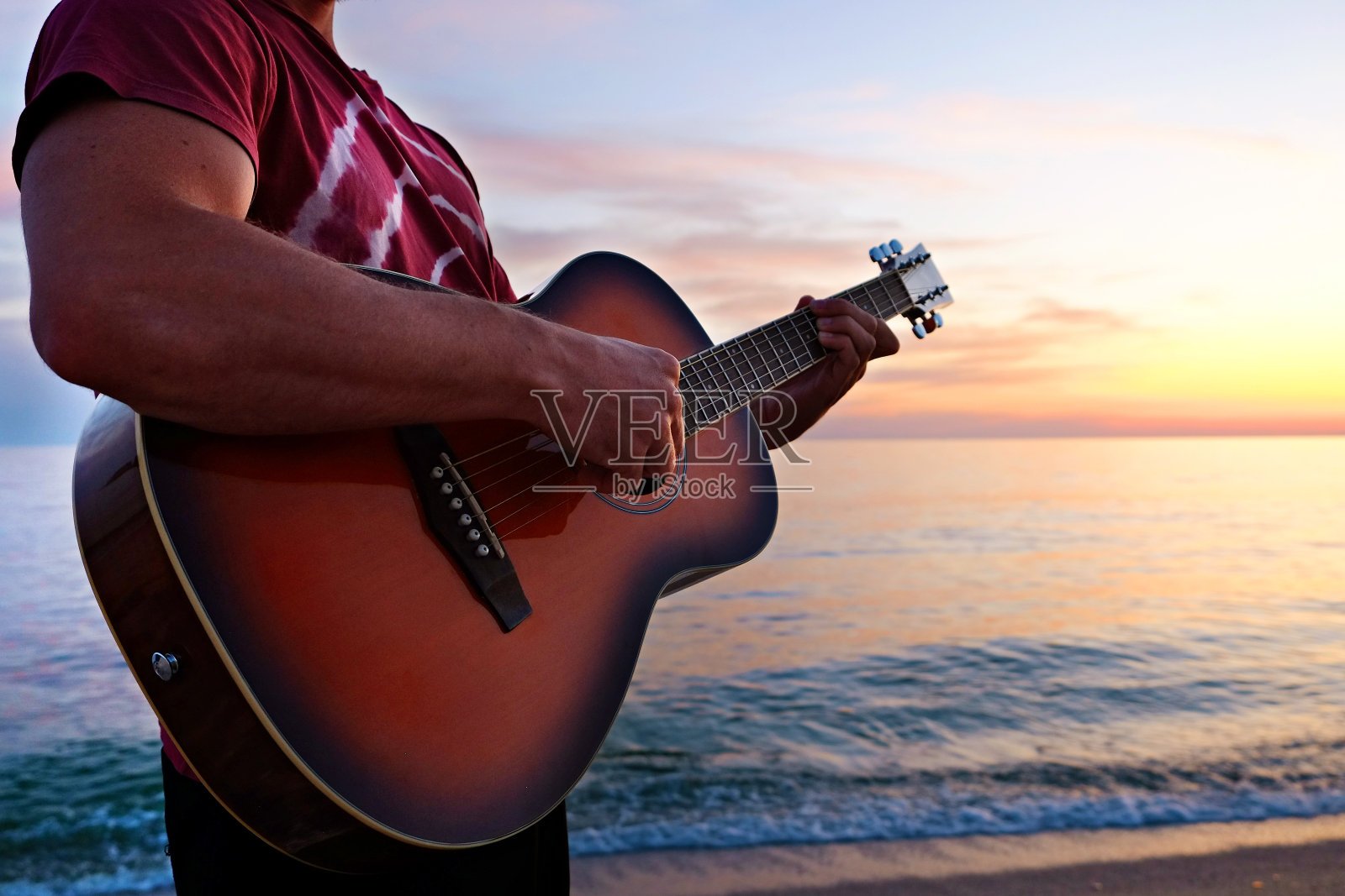 年轻人穿着紫色的扎染t恤，在美丽的日落时分，在海滩上弹无畏客厅的原声吉他。适合吉他手与日落的乐器在海边。背景，拷贝空间，关闭照片摄影图片
