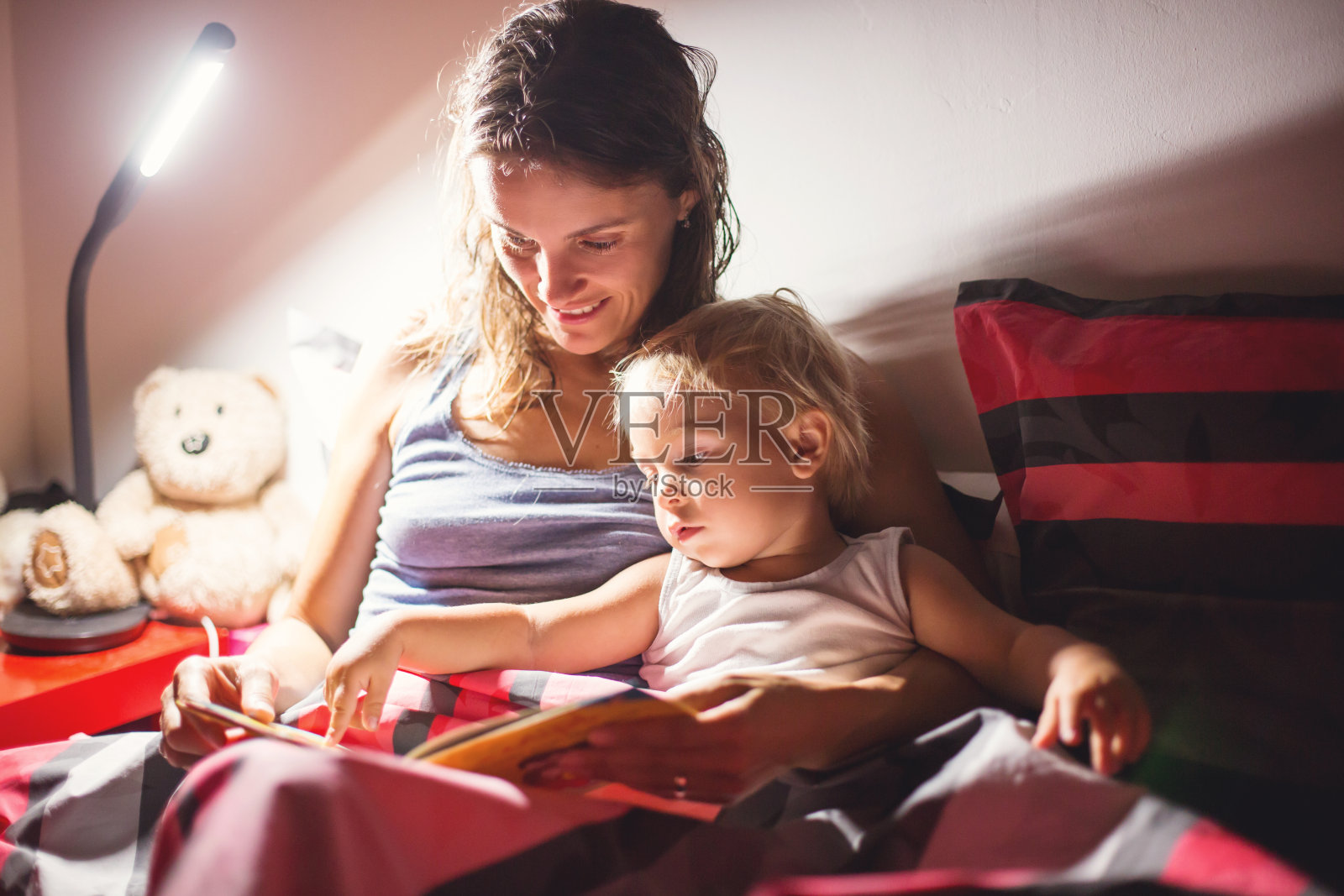 母亲晚上在家给蹒跚学步的儿子读书时，床头灯开了照片摄影图片