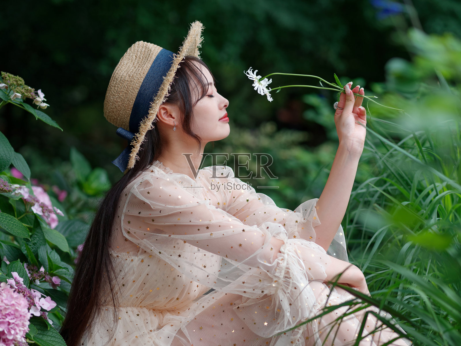 美丽的中国女孩穿着礼服，戴着帽子坐在花园里，闭着眼睛，手里拿着一朵白色的野花，享受着她在夏天的闲暇时光，侧视图。照片摄影图片