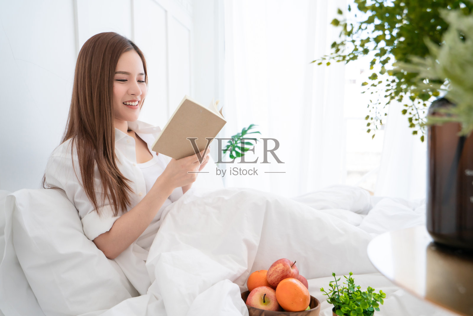 美丽的亚洲女性白衣长发在白色的床上以蛤蜊和欢快的白色卧室室内为背景读书照片摄影图片