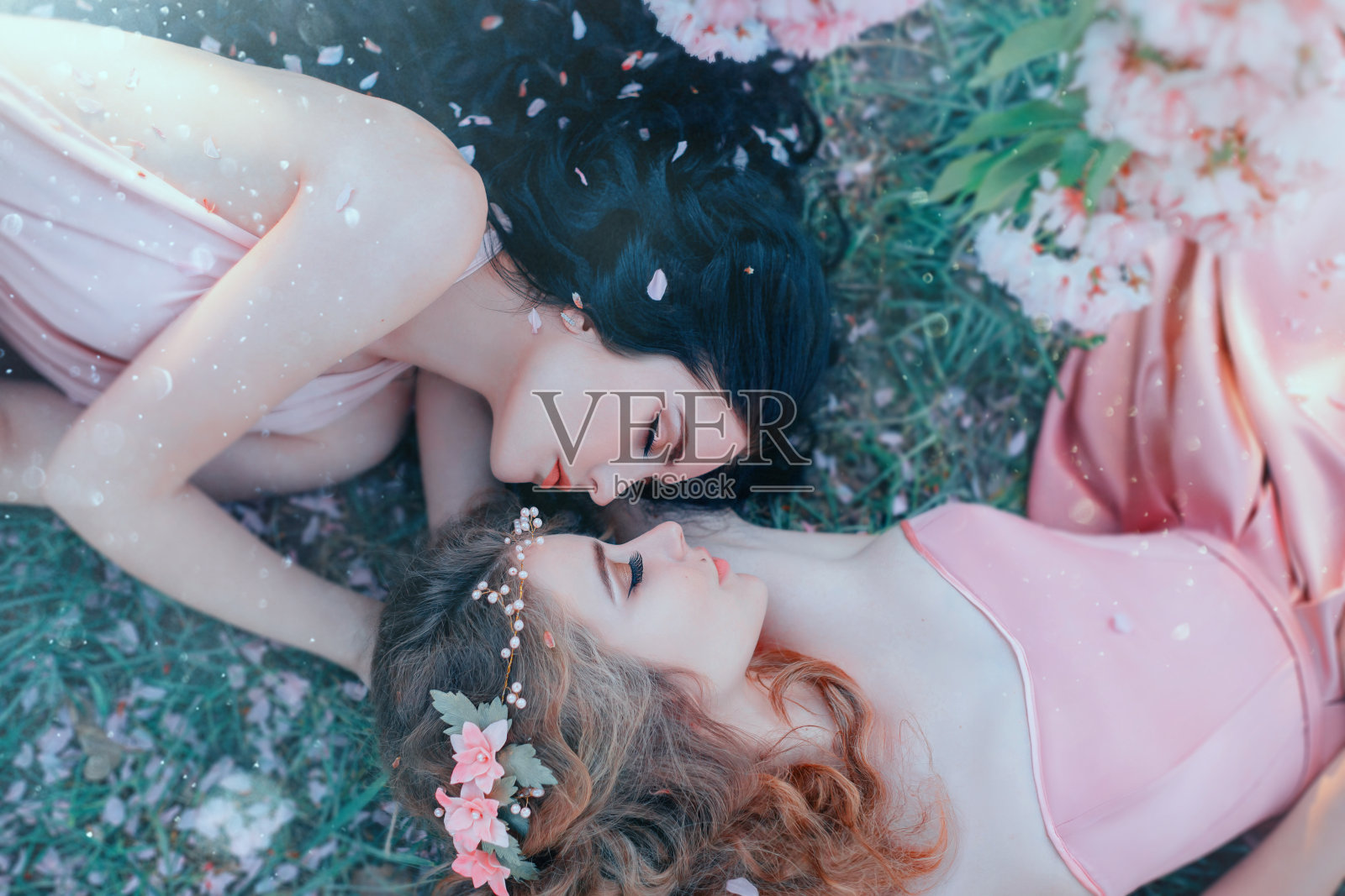 两名年轻女子躺在春天花园的草地上拥抱，华丽丽的长卷发上撒满了鲜花和玫瑰花瓣。迷人的脸庞，柔和的妆容。不同的姐妹。天然化妆品的概念照片摄影图片