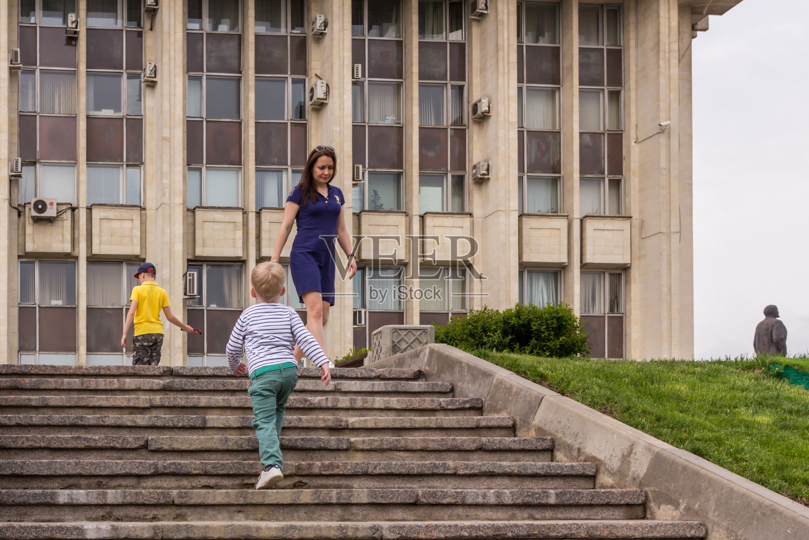一位母亲和她的儿子们在现代建筑背景的石头楼梯上休息。女人和男孩在公园里玩。照片摄影图片