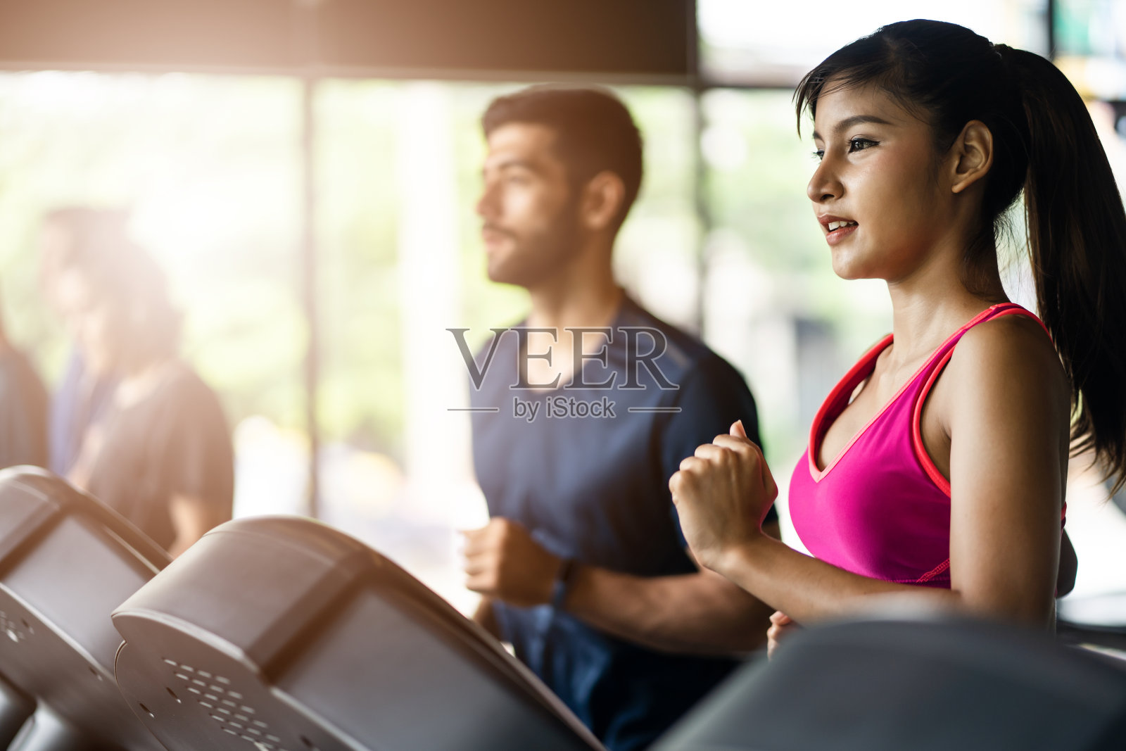 亚洲健康美丽的女运动员在运动服慢跑在跑步机上或跑步机与高加索运动男子在健身房或健身。女人面带微笑，快乐地看着前面。为了健康而锻炼。照片摄影图片