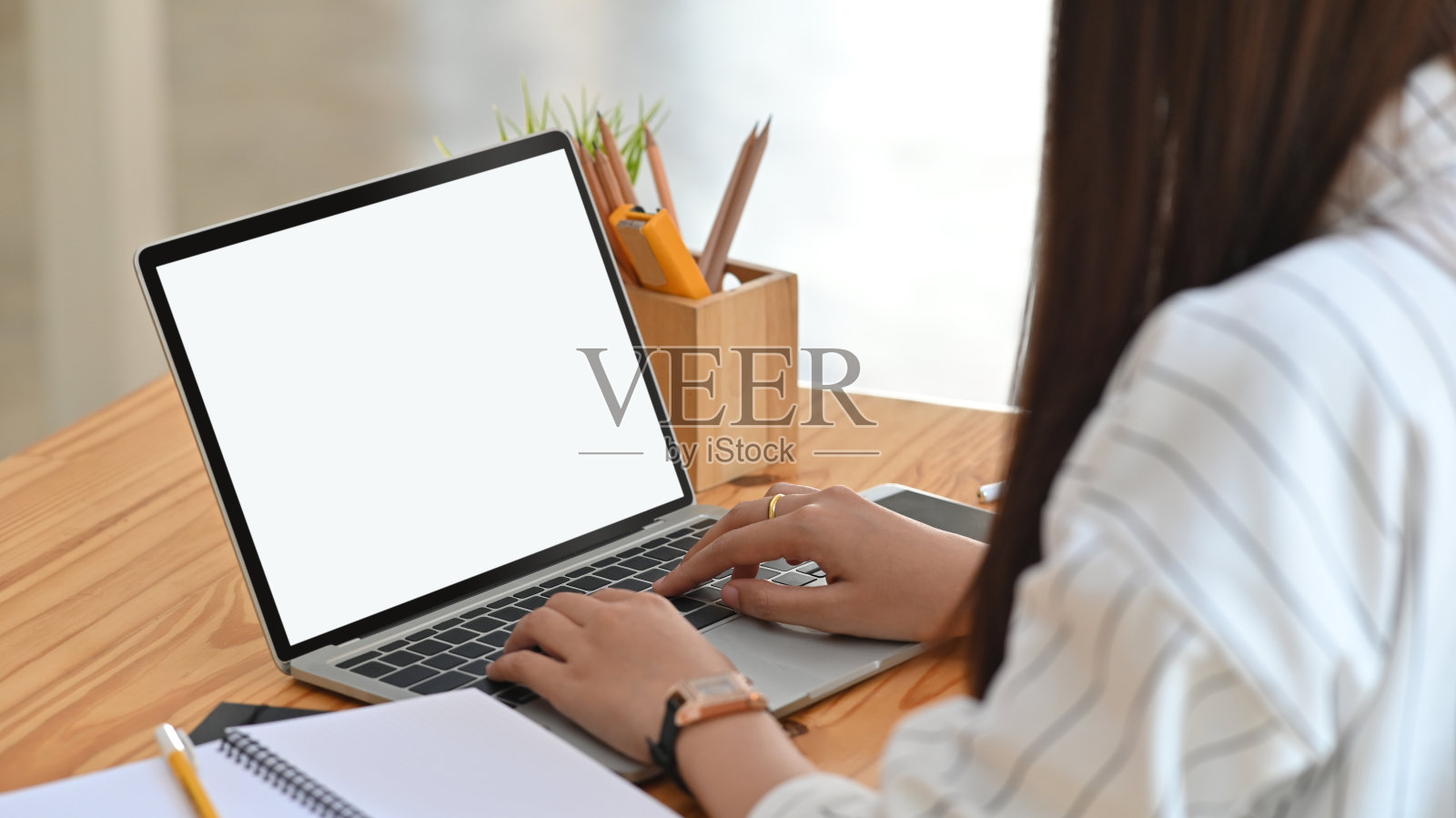 这是一名年轻女子在笔记本电脑上打字时的侧影。盆栽植物，黑屏智能手机，笔筒，木桌笔记。照片摄影图片