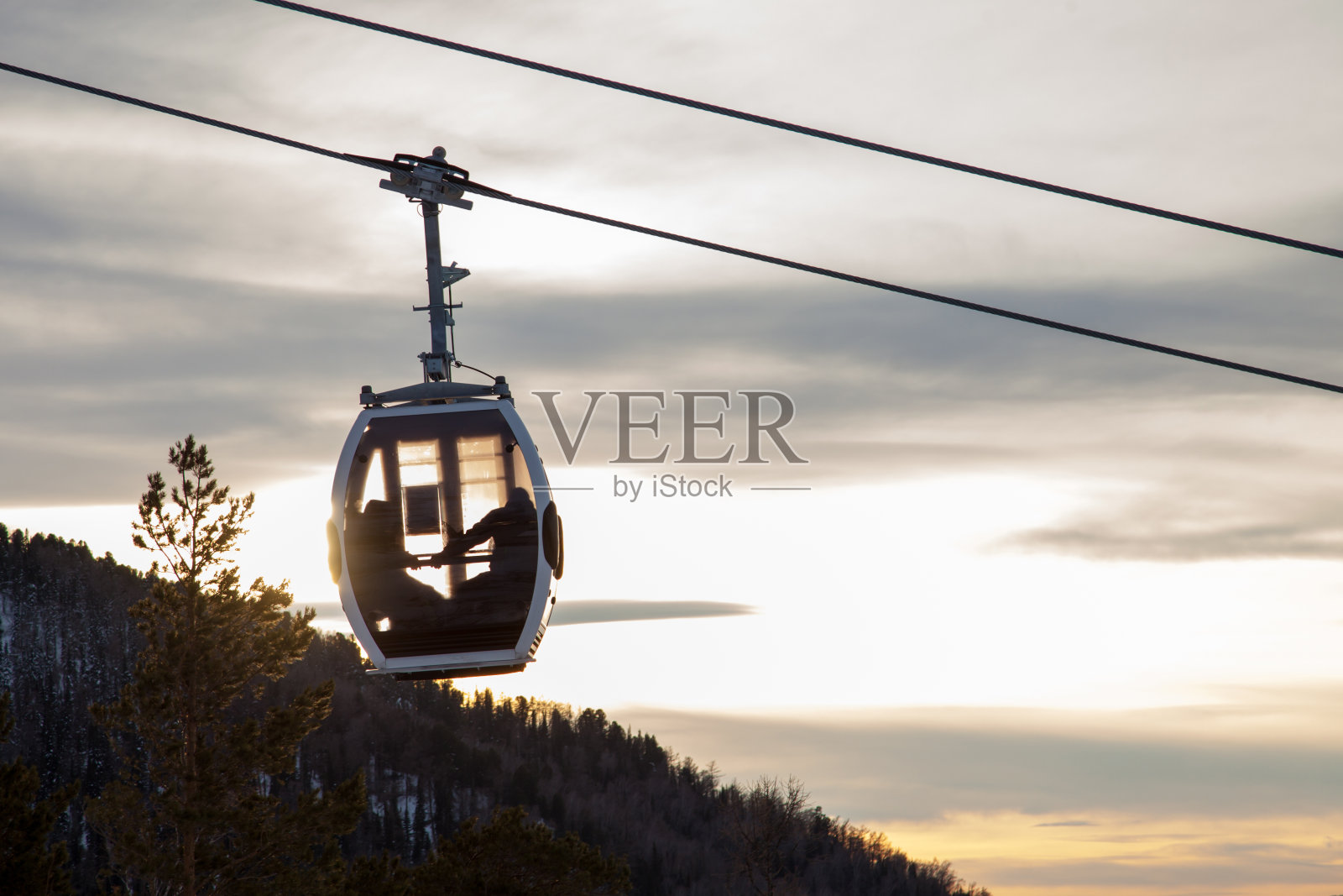 这是一种带有悬挂在缆索上的小亭子的缆车，情侣们坐在上面，背景是群山、树木和夕阳。滑雪胜地和滑雪板。照片摄影图片