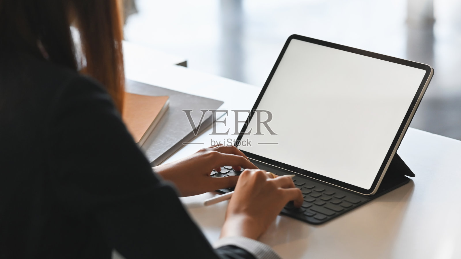 这张照片拍摄的是一位年轻的女商人坐在现代办公桌旁，用白色的空白屏幕敲击平板电脑键盘。照片摄影图片