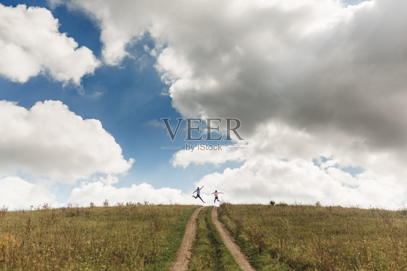 快乐活跃的夫妇跳跃在绿色的田野与蓝天与云彩。暑假的概念。远处一对夫妇的照片照片摄影图片